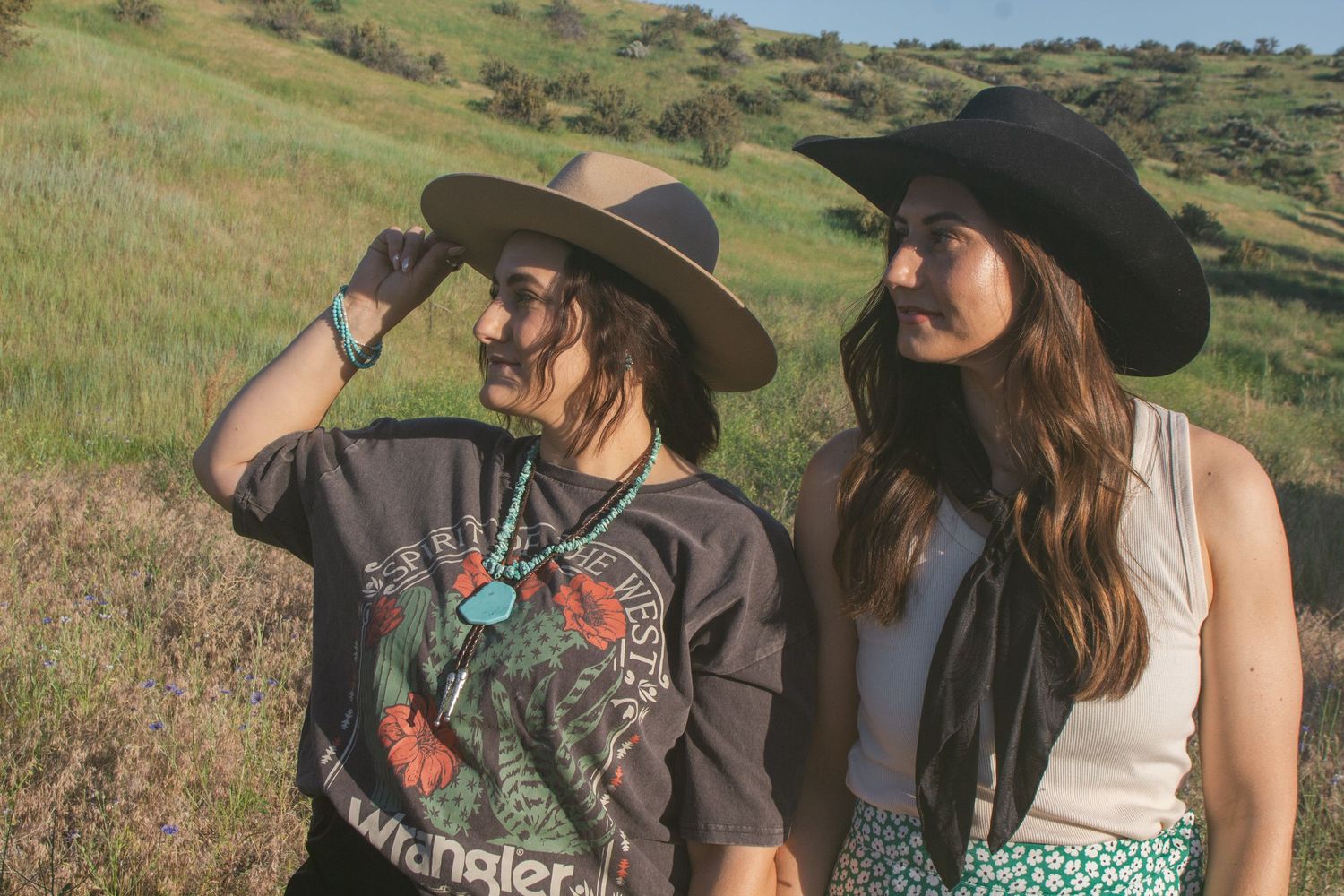 Two women in cowboy-style hats stand side by side on a green hillside, one shielding her eyes from the sun.