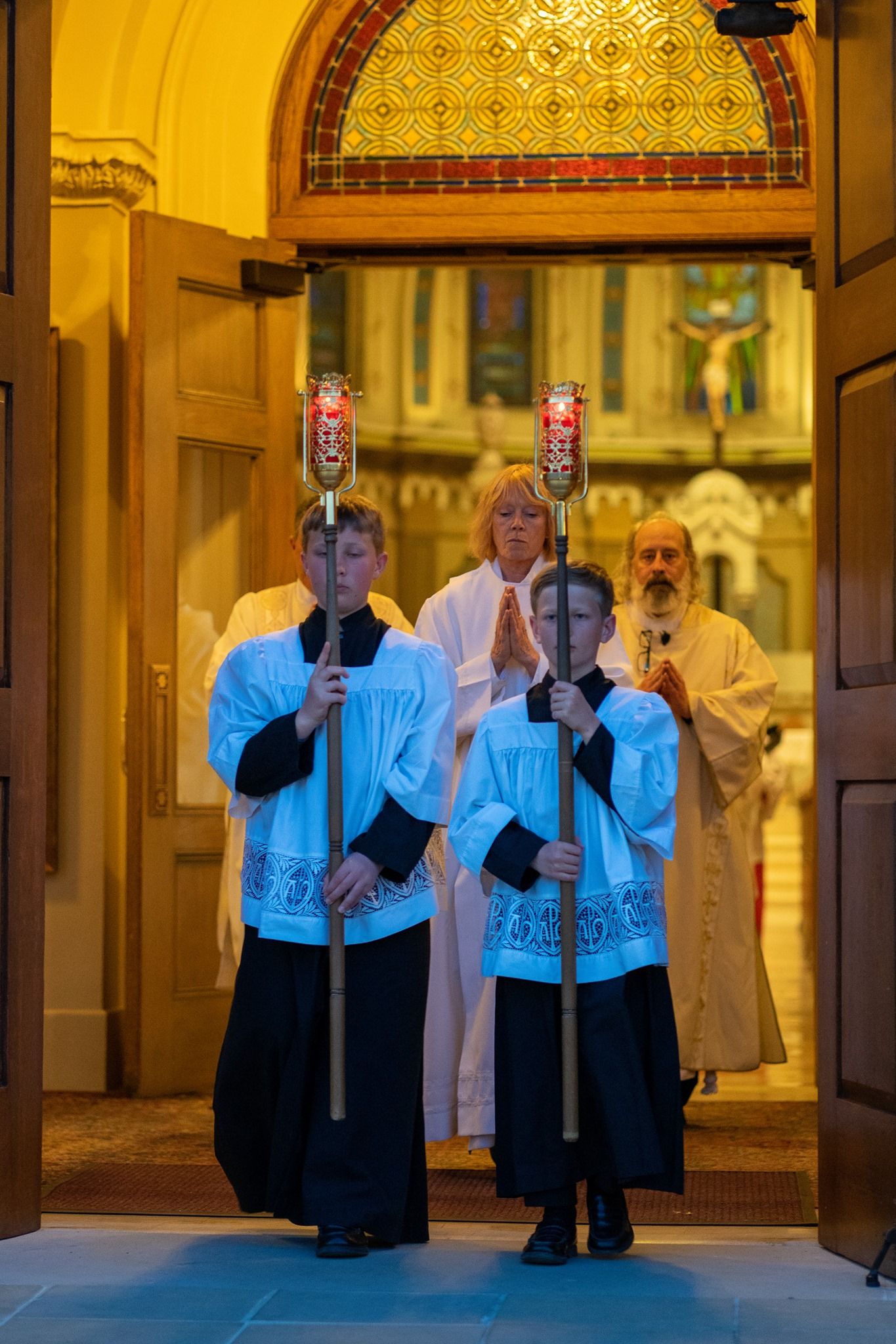 Two altar servers in white surplices carry tall candle torches through church doors, followed by clergy in a solemn procession.