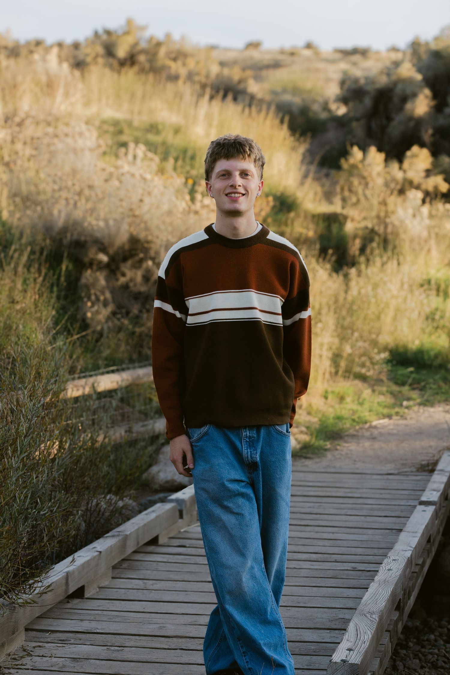 Smiling young man in a brown striped sweater and jeans stands on a wooden footbridge near golden grasses.