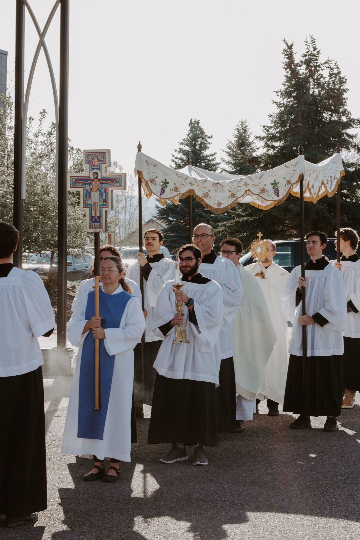 Altar servers and clergy in white robes walk in a Eucharistic procession under a canopy, carrying a processional cross outdoors.