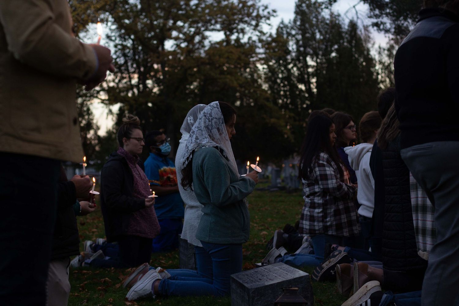 A group of people kneel on the grass holding candles during an outdoor evening prayer vigil at dusk.