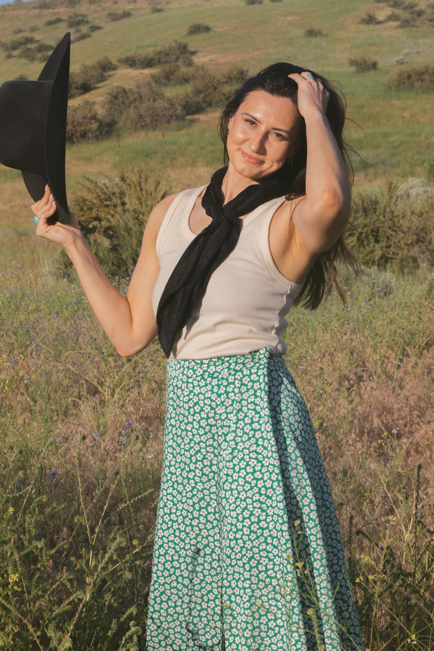Smiling woman in a cream tank top, black scarf and green floral skirt holds a black cowboy hat in a sunny grassy field.