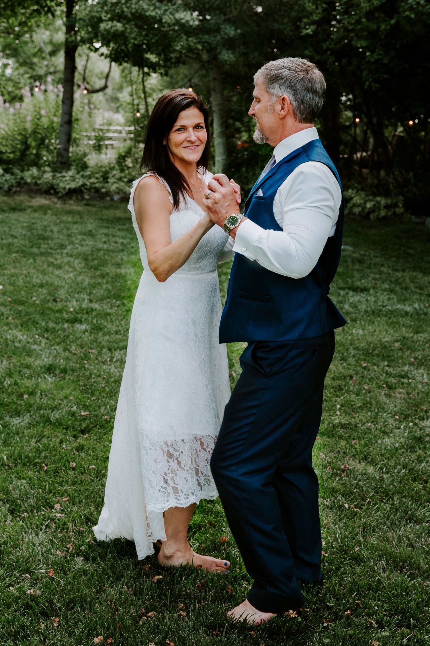A smiling barefoot bride in a white lace dress dances with her groom in a navy vest on a green lawn.