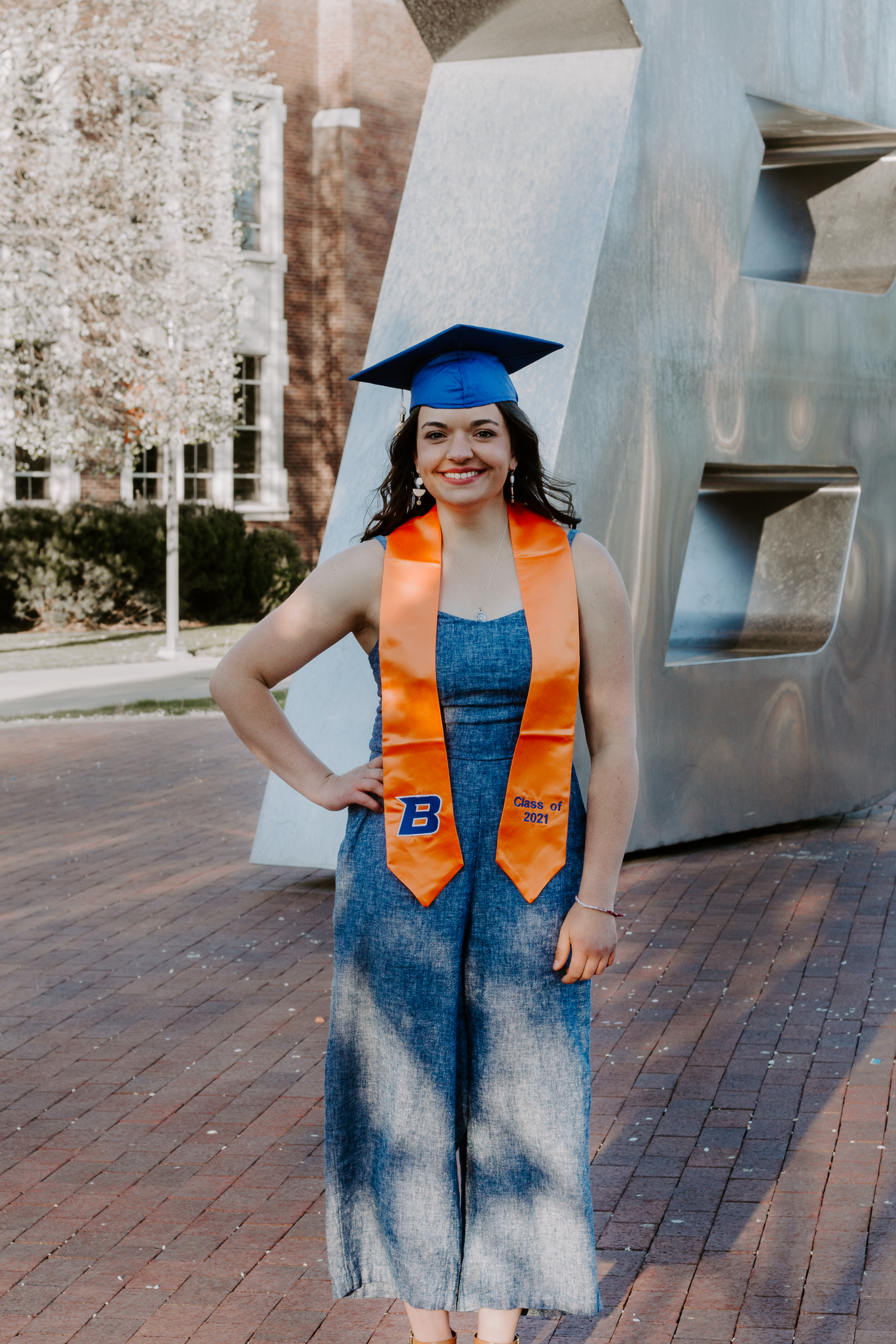 Smiling graduate in a blue cap, orange Boise State stole and denim jumpsuit poses with a hand on her hip beside a metal sculpture.