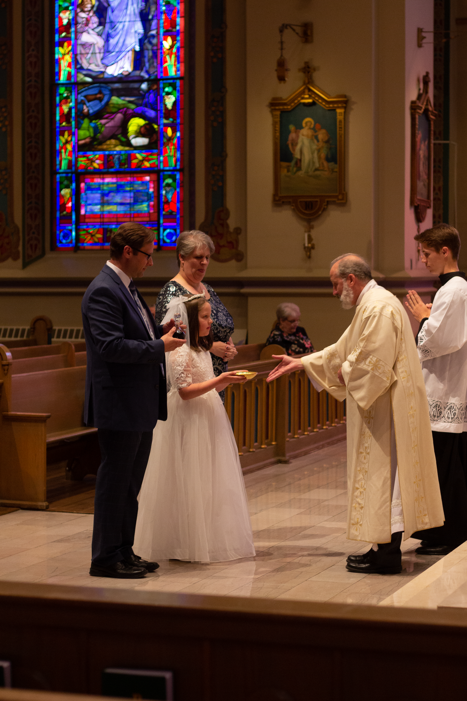 A girl in a white dress receives a communion offering from a priest as her parents and an altar server look on.