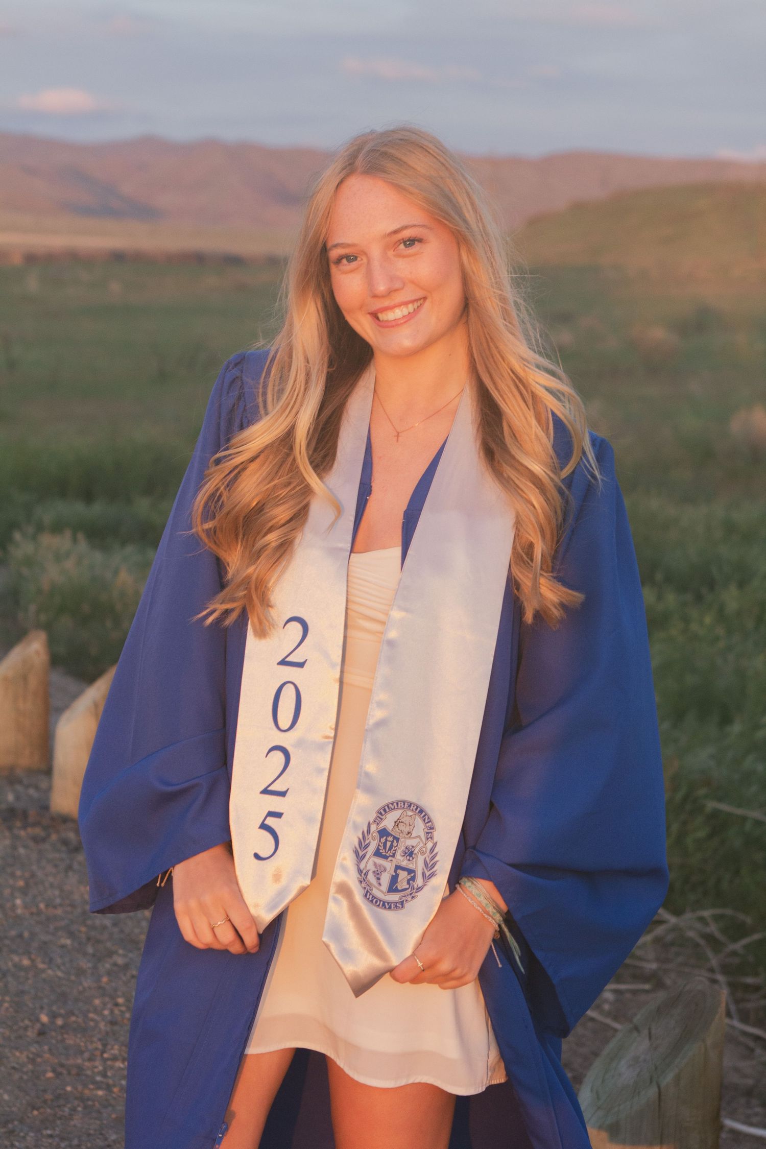 Smiling graduate in a blue gown and white 2025 stole stands in a grassy field with hills glowing at sunset.