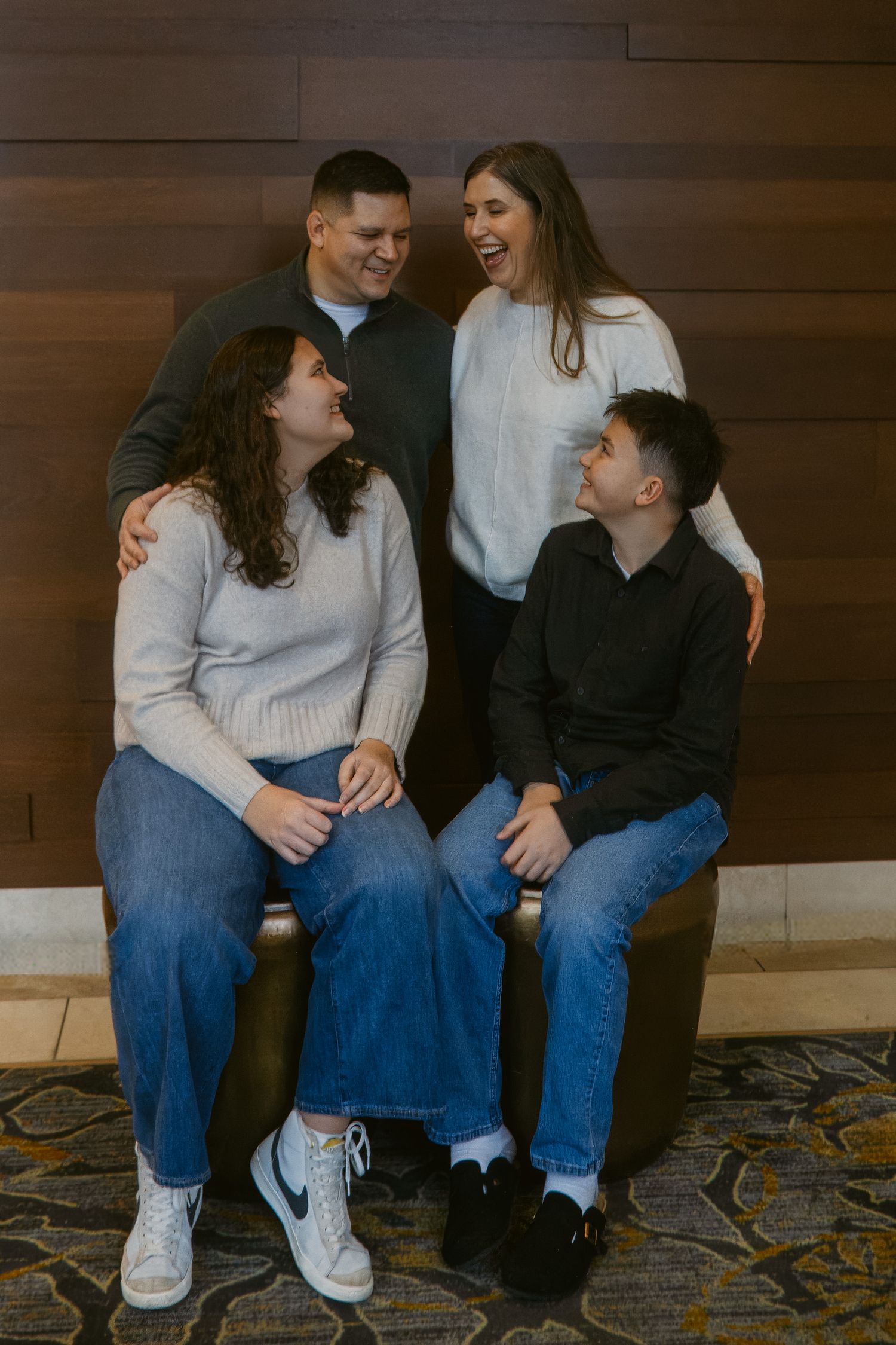 Family of four laughs together in a cozy lobby, with parents standing behind two teenagers seated on a bench.