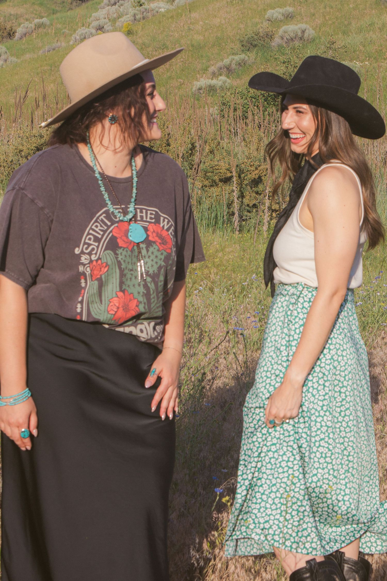 Two laughing women in hats and western boho outfits chat in a grassy field, one wearing a turquoise necklace.