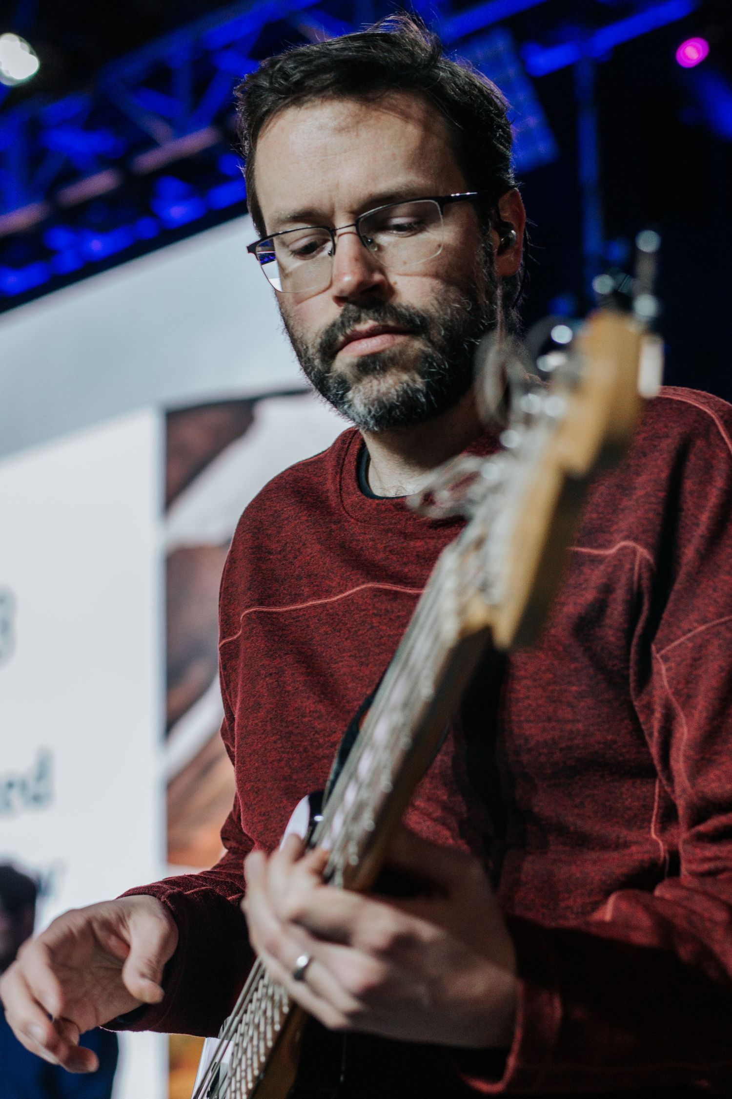 A bearded man with glasses and a red sweater plays bass guitar on a stage with blue lighting.