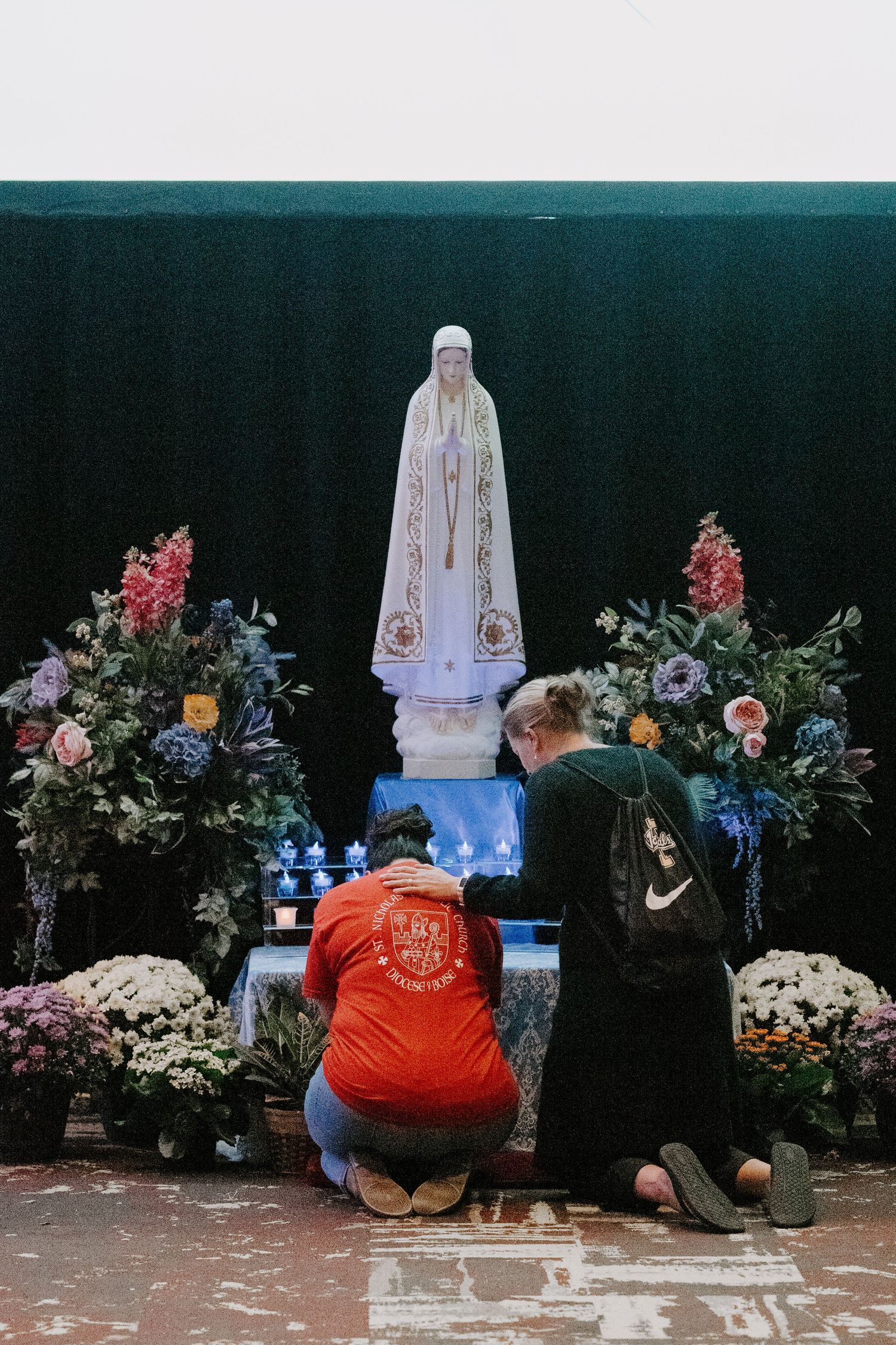 Two people kneel in prayer before a white Our Lady of Fatima statue surrounded by colorful flowers and blue candles.