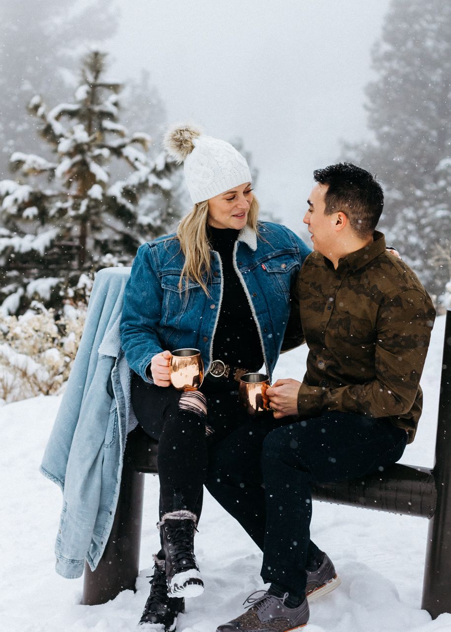 A couple in winter clothing sit on a bench holding copper mugs and smiling at each other in a snowy forest.