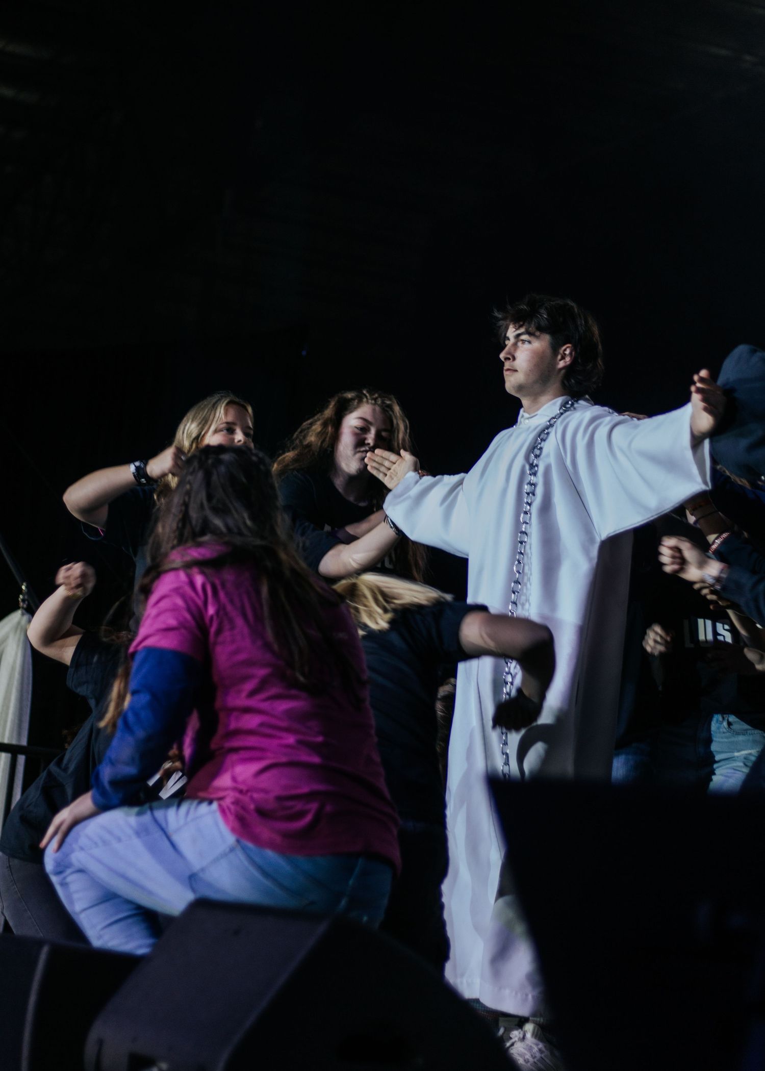 A young man in a white robe extends his arms toward a group of teens gathered close to the stage during a worship moment.