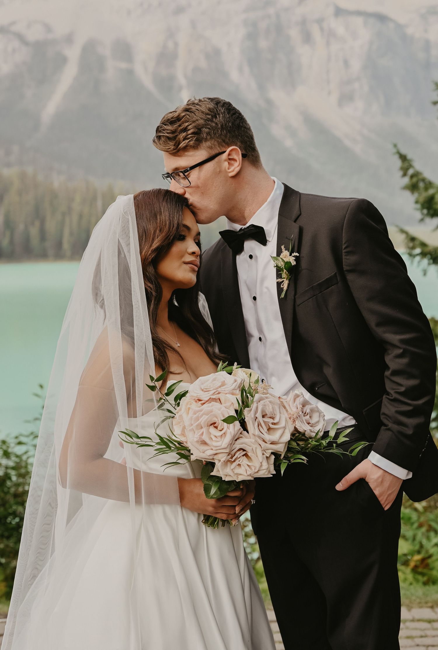 A romantic wedding couple embraces by a turquoise lake with majestic mountains in the background.