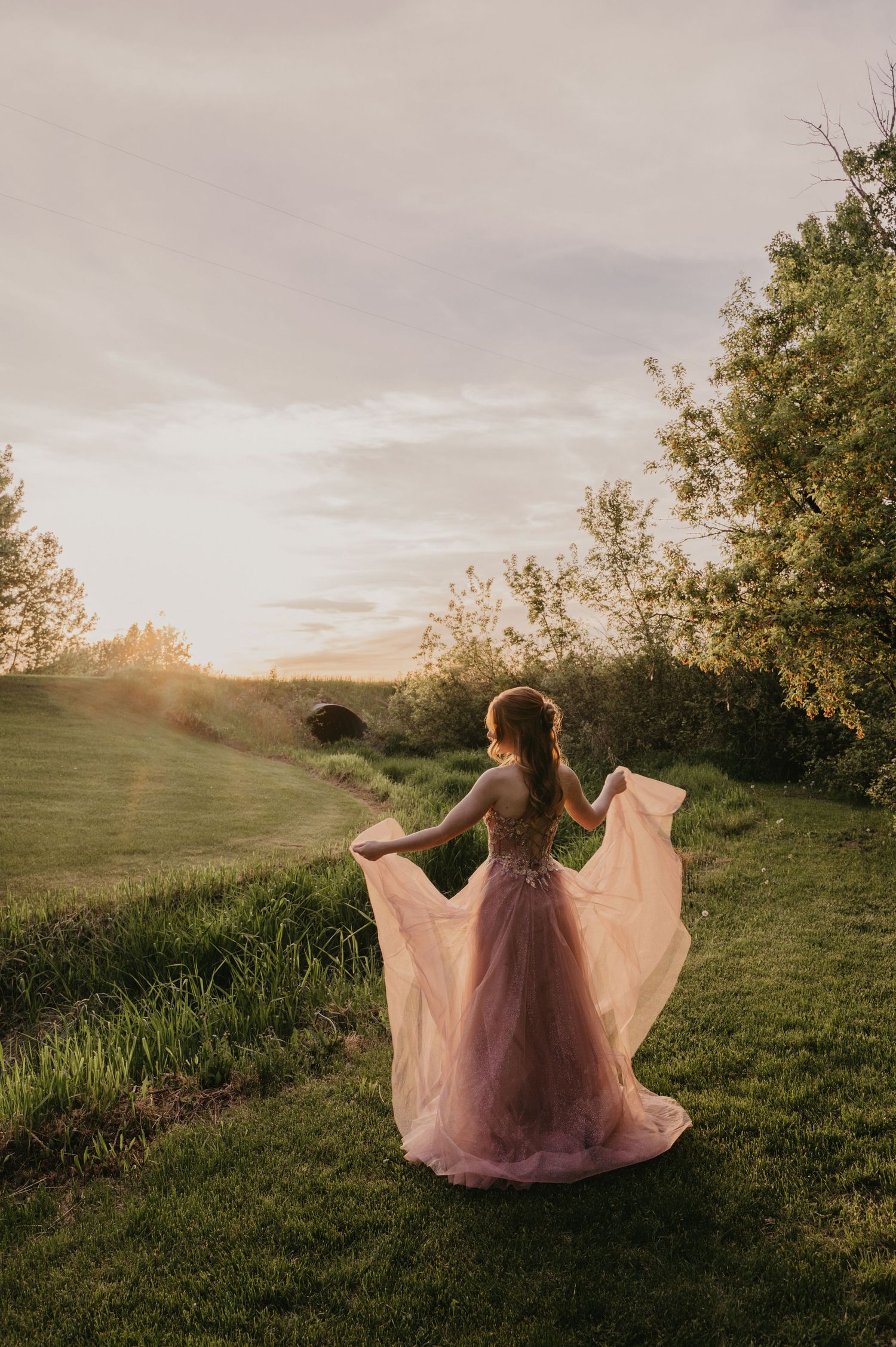 Figure in flowing pink dress twirls gracefully on grassy hillside at sunset creating ethereal silhouette against golden sky.