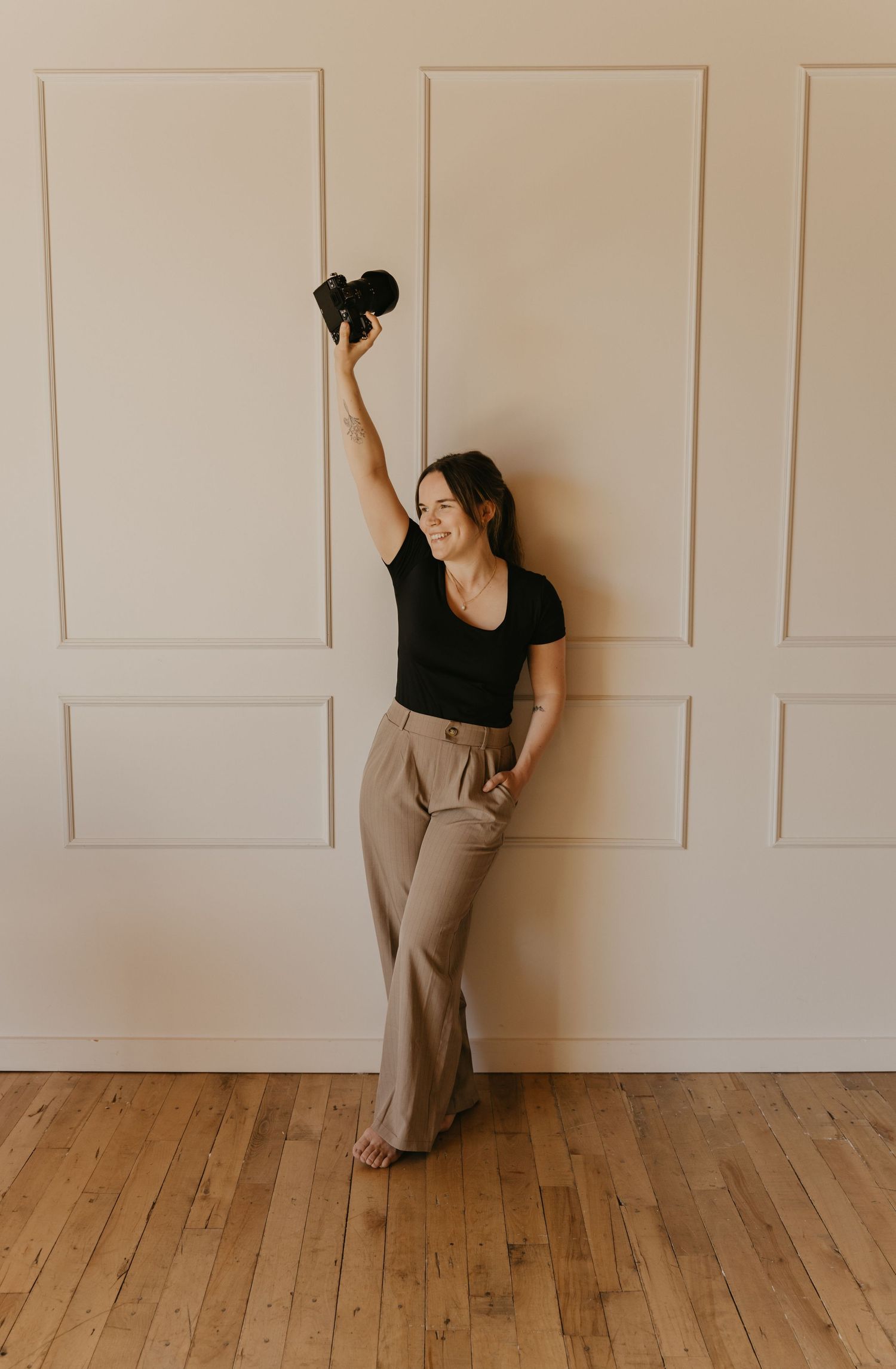 A person in black top and tan pants holds up dumbbells while standing against a white paneled wall and wooden floor.