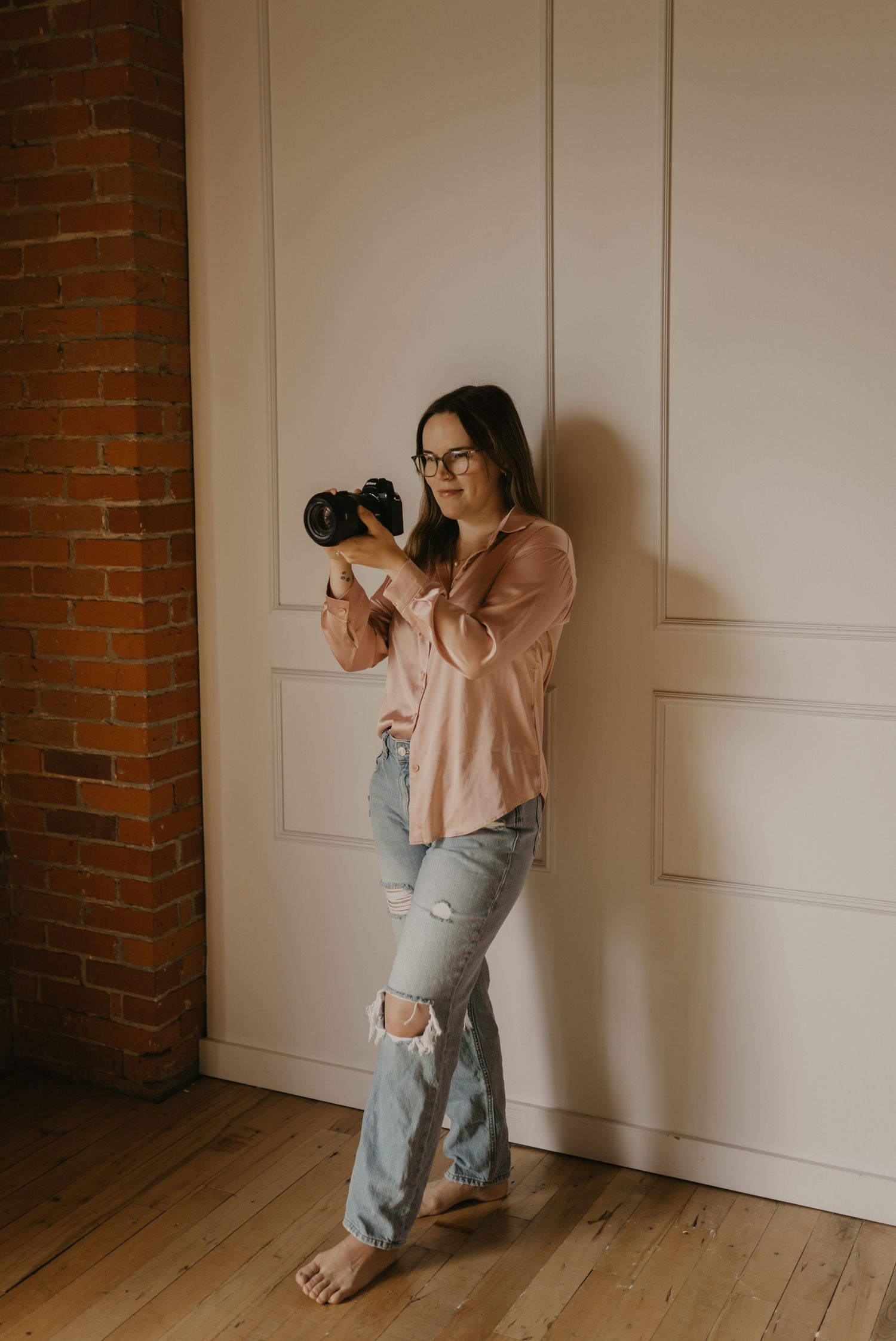 A photographer in ripped jeans and peach sweater poses with camera against white wall and exposed brick background.