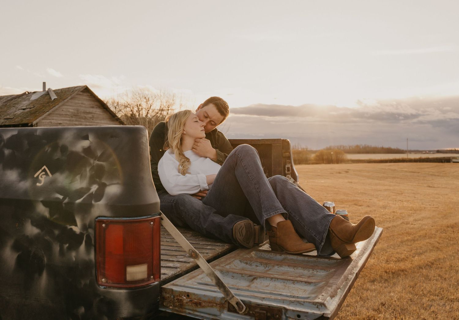 Couple relaxes in pickup truck bed watching sunset over golden wheat fields during summer evening.