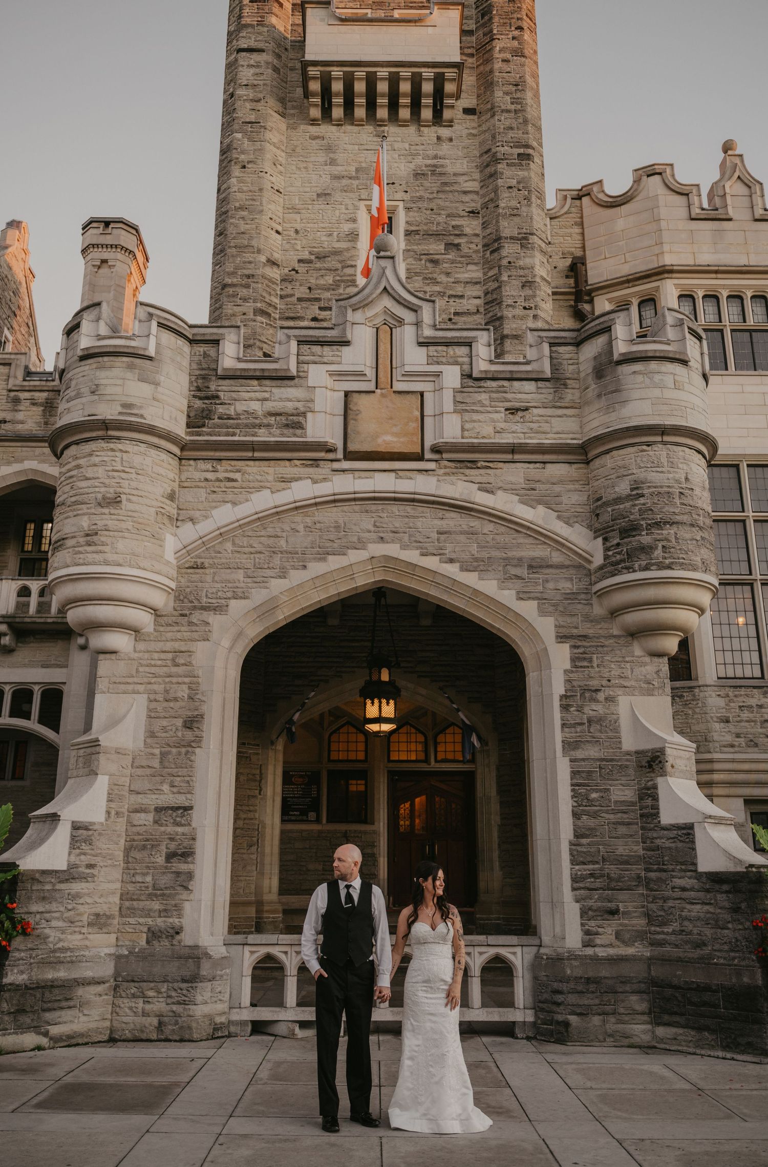 A wedding couple stands in front of a grand gothic stone castle entrance with turrets and arched doorway at dusk.