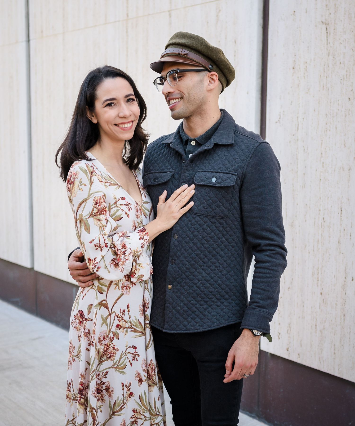 A couple poses together against a white wall, with her wearing a floral dress and him in a quilted jacket and cap.