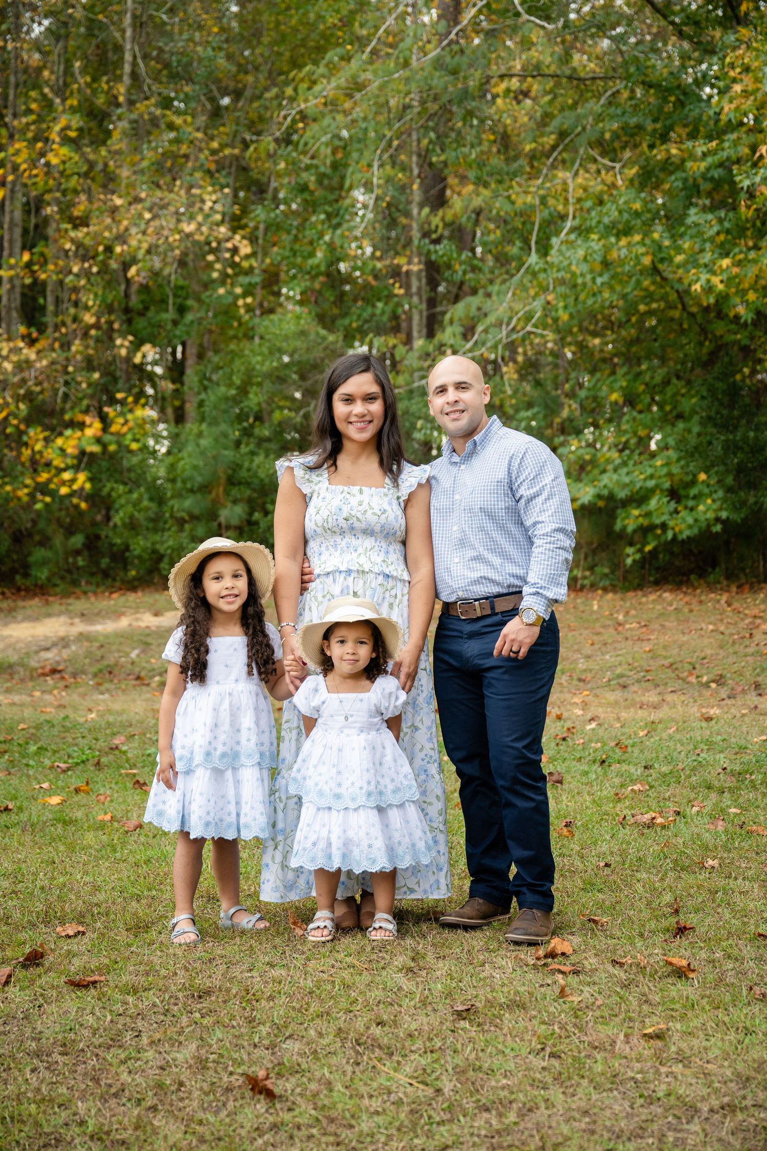 A family wearing coordinating blue and white outfits poses together on grass with autumn trees in the background.
