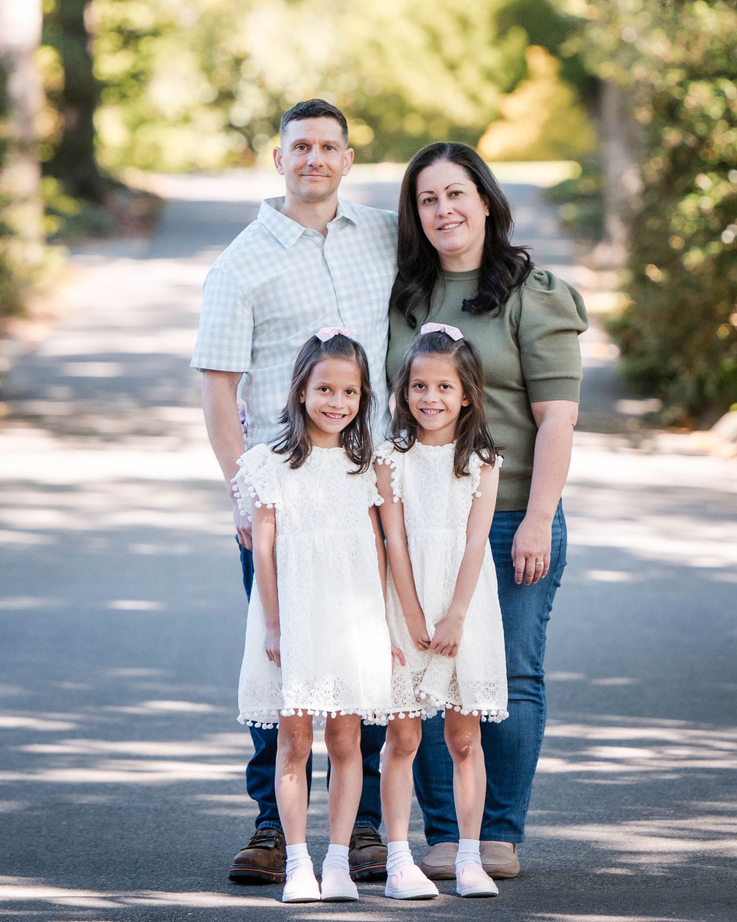 Family of four poses outdoors on a tree-lined path, parents stand behind two young girls in white dresses.