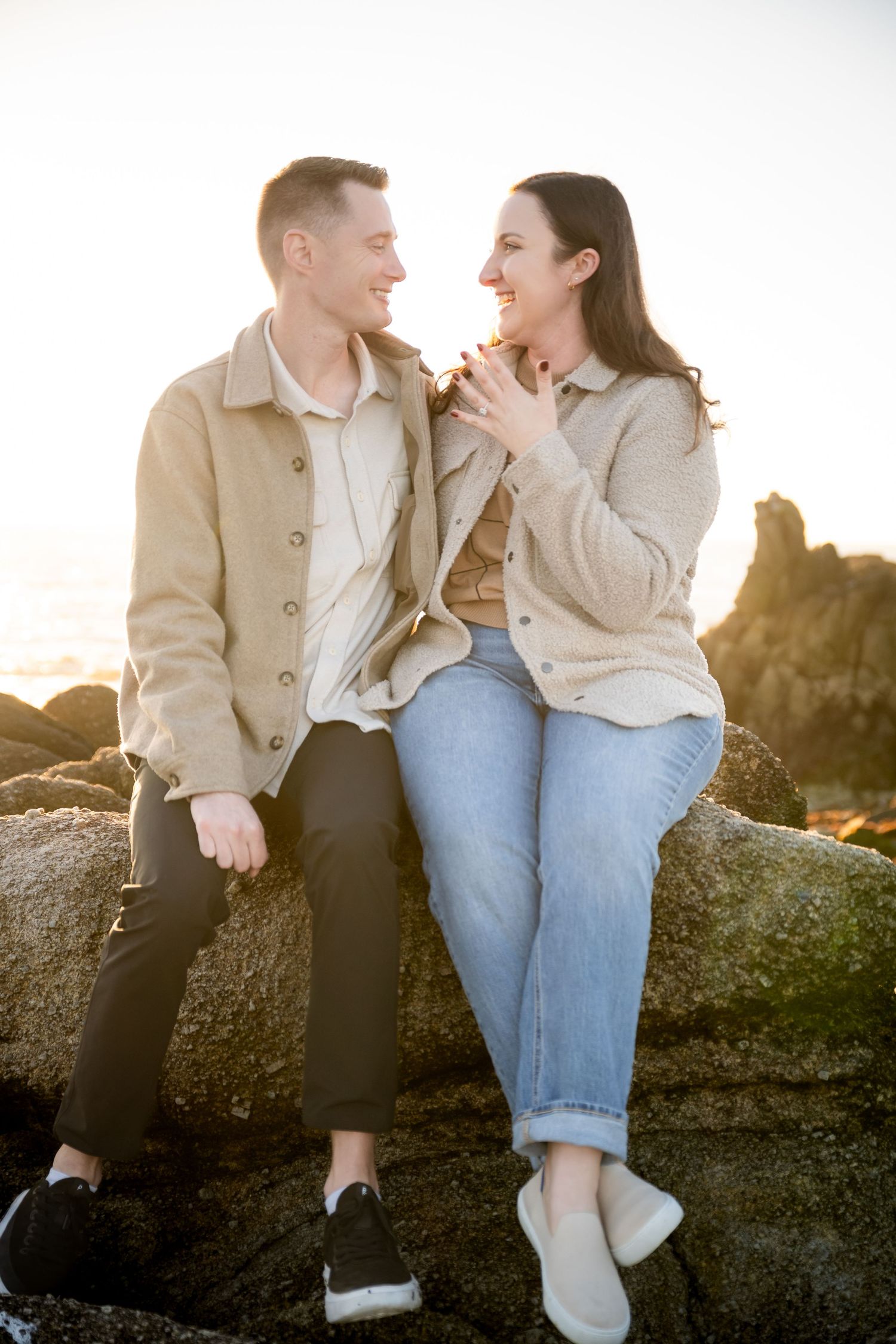 Couple sitting on rocks at sunset, smiling at each other, wearing casual beige jackets and jeans.