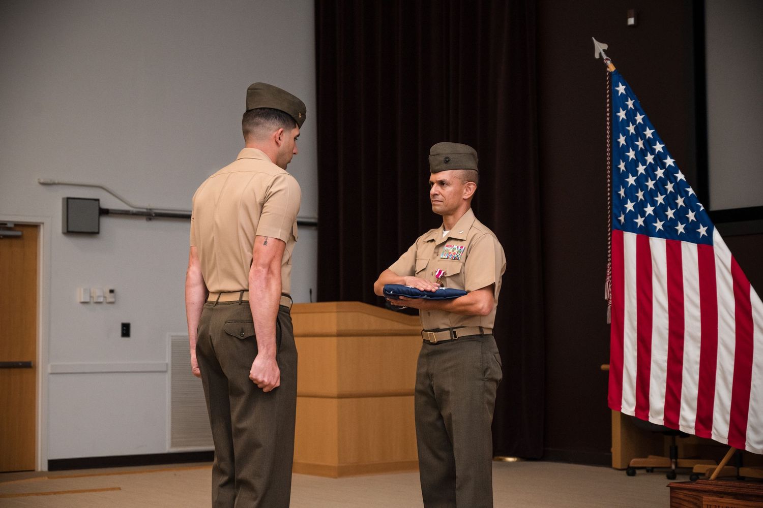 Military personnel stand in formation during a ceremony with an American flag displayed on stage.
