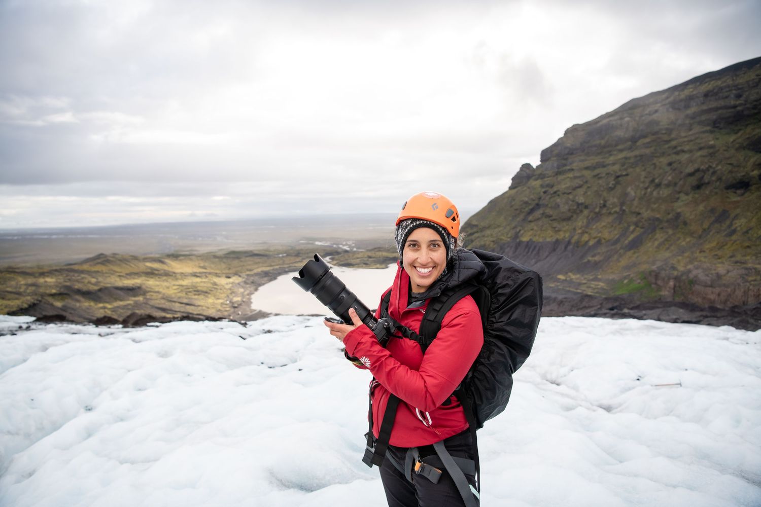 A person in a red jacket stands on a snowy mountain peak with dramatic cloudy skies in the background.