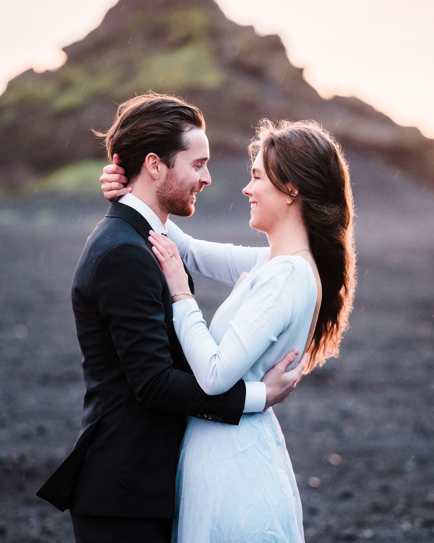 A romantic couple in formal attire share intimate moments on a rocky beach at sunset.
