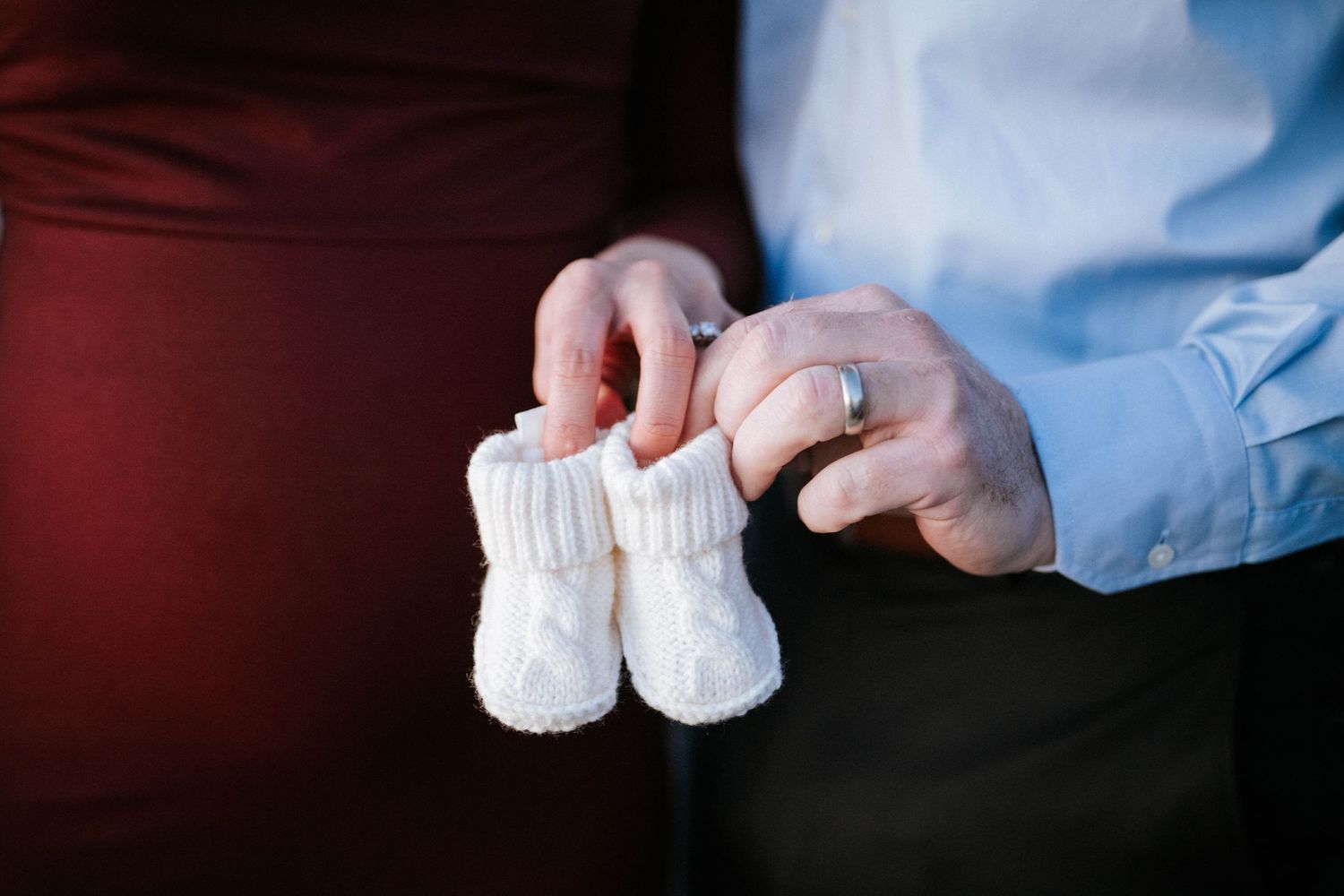 Couple holding tiny white knitted baby booties, announcing pregnancy, with wedding ring visible on hand.