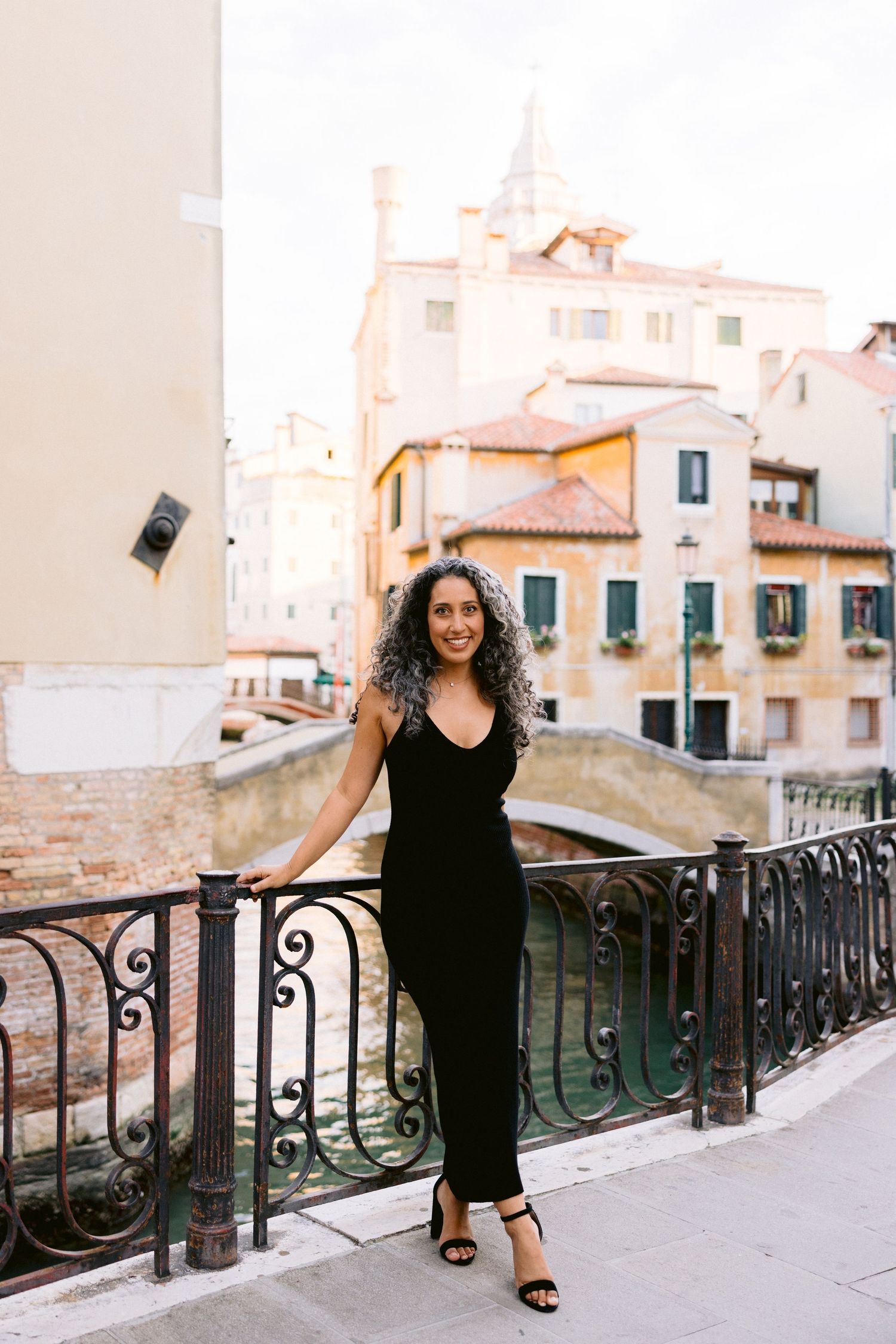 A person in a black dress poses along an ornate iron railing on a Mediterranean balcony with white buildings in view.