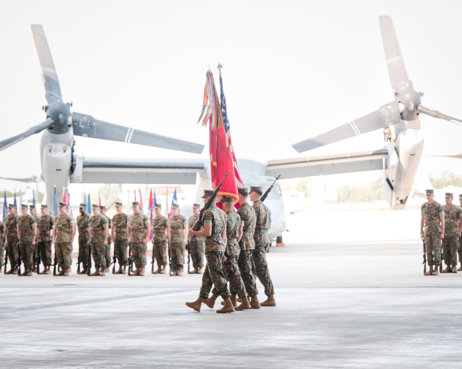 Military personnel stand in formation on an airfield with aircraft visible in the background.