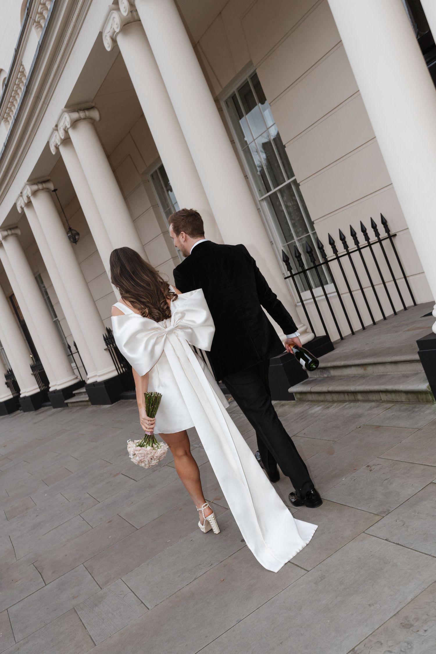 A couple in wedding attire walking together under classical white columns of a historic building.