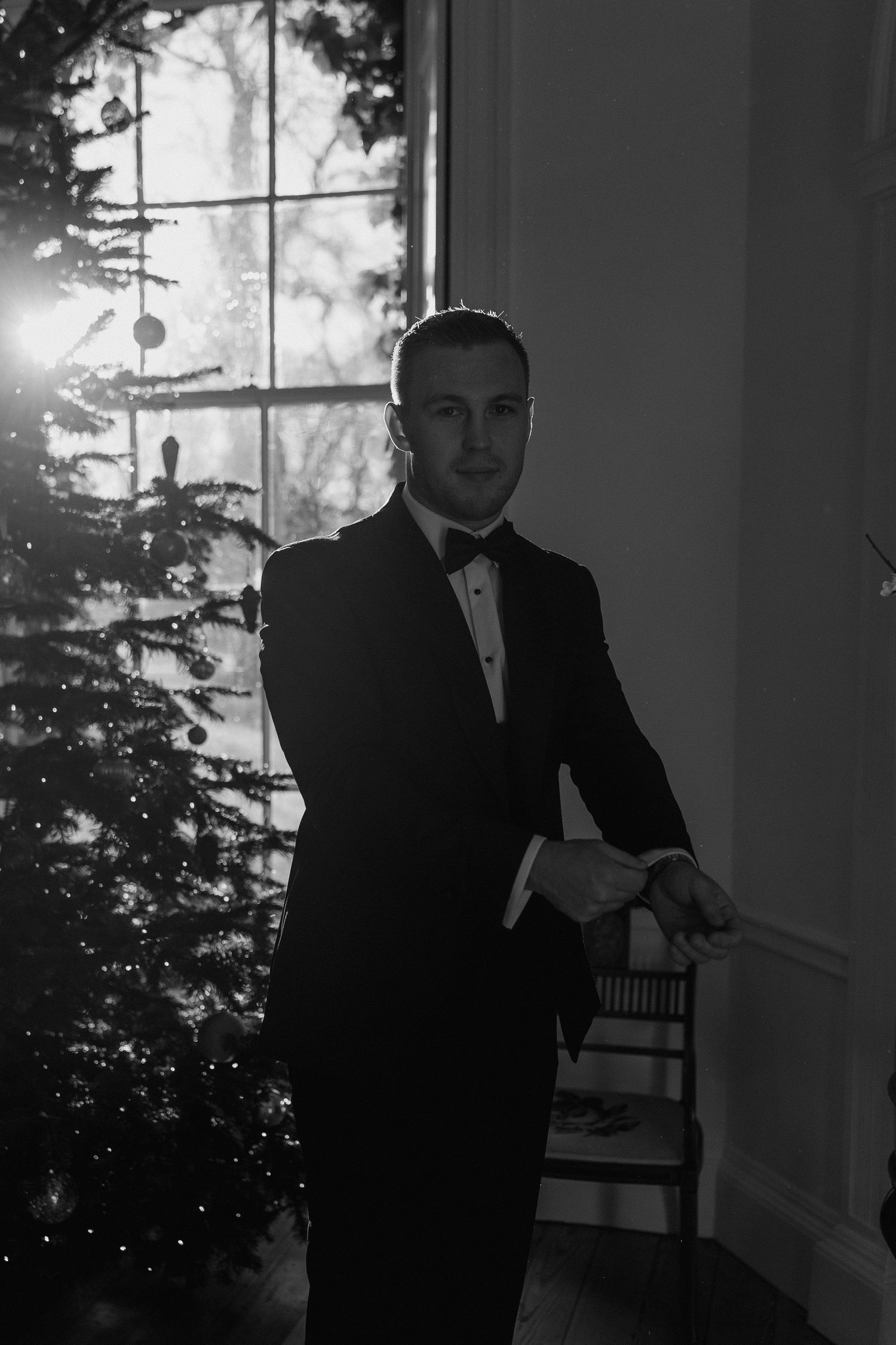 Groom in classic black tuxedo poses by window with dramatic backlighting at sunset.