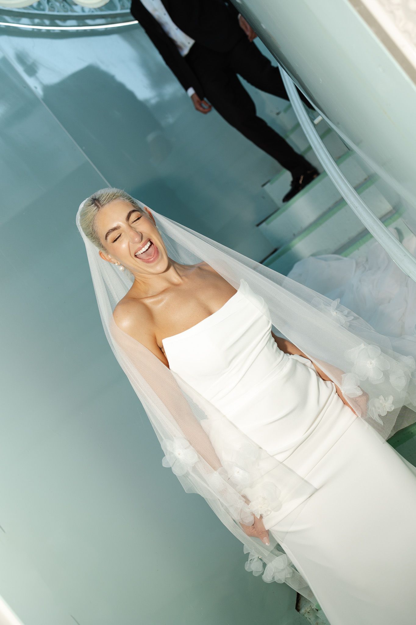 A bride in elegant white strapless dress and veil smiles radiantly in front of staircase.