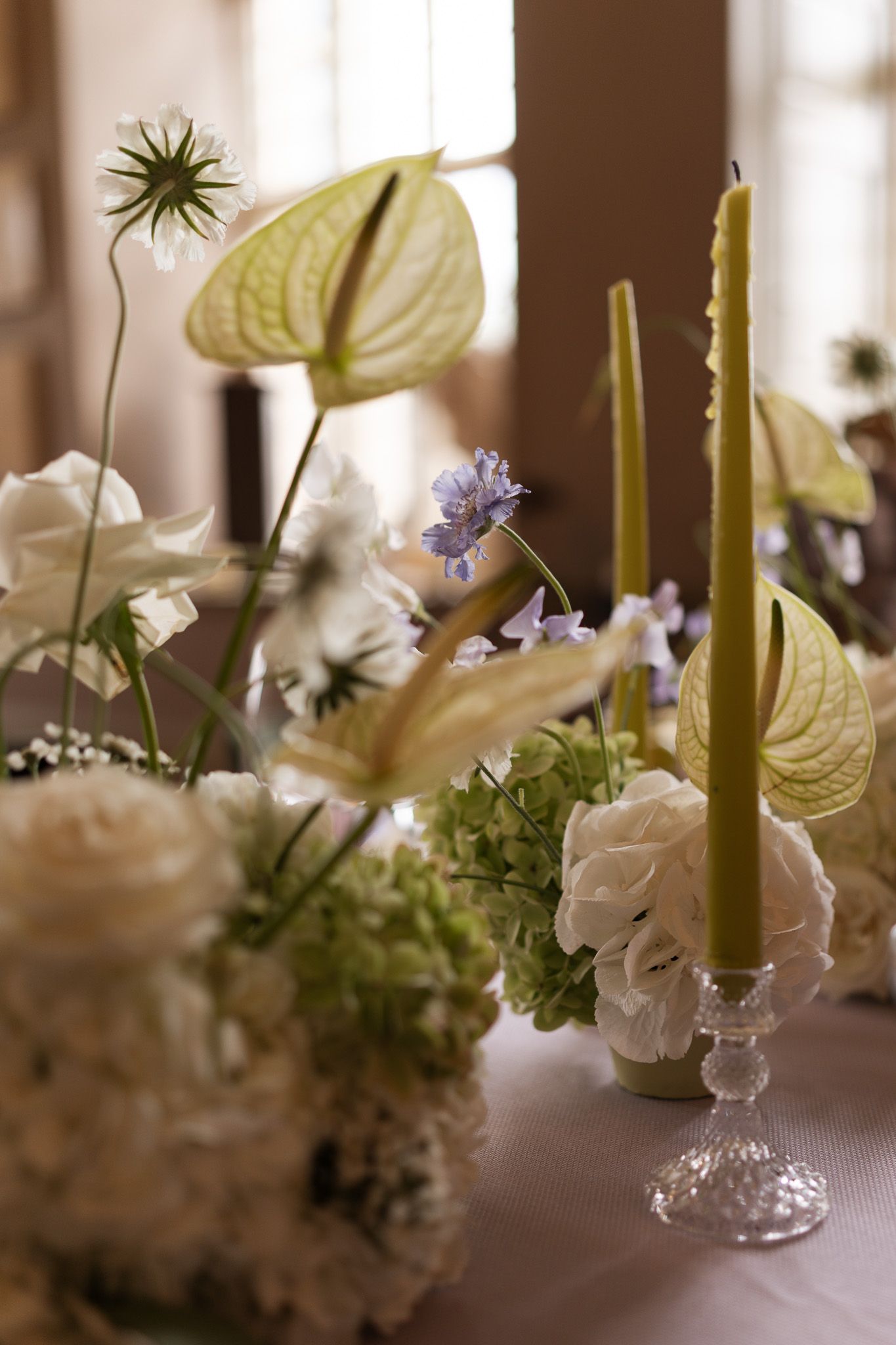 Elegant floral arrangement with anthurium lilies, delicate blue flowers, and white blooms beside a yellow taper candle in glass holder.