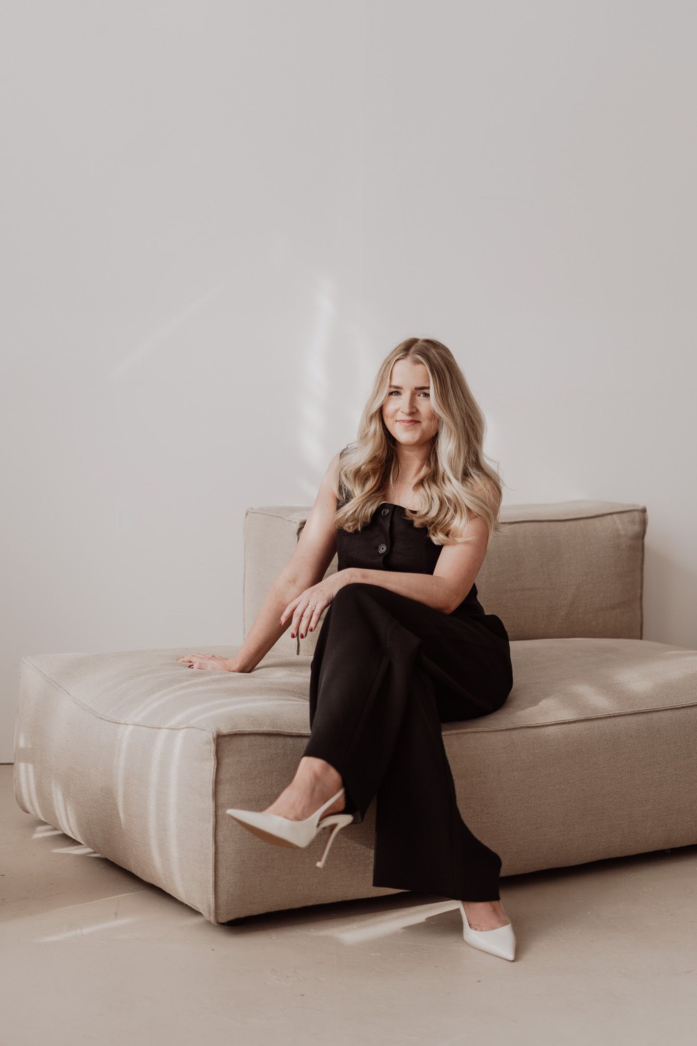 A professional portrait of someone sitting on a beige couch wearing a black dress and white heels.