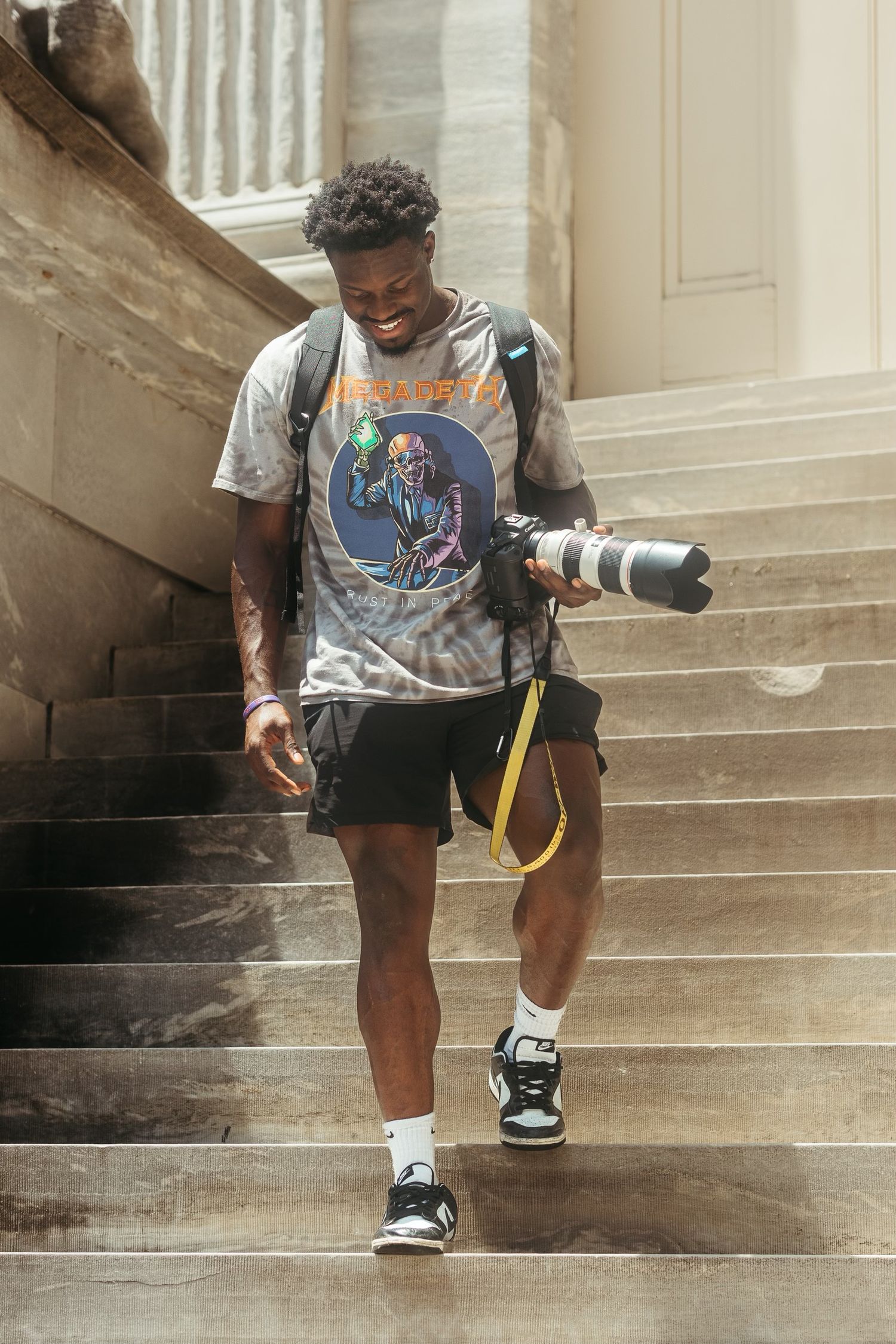 Person sitting on concrete steps wearing a graphic t-shirt and shorts holds a professional camera.