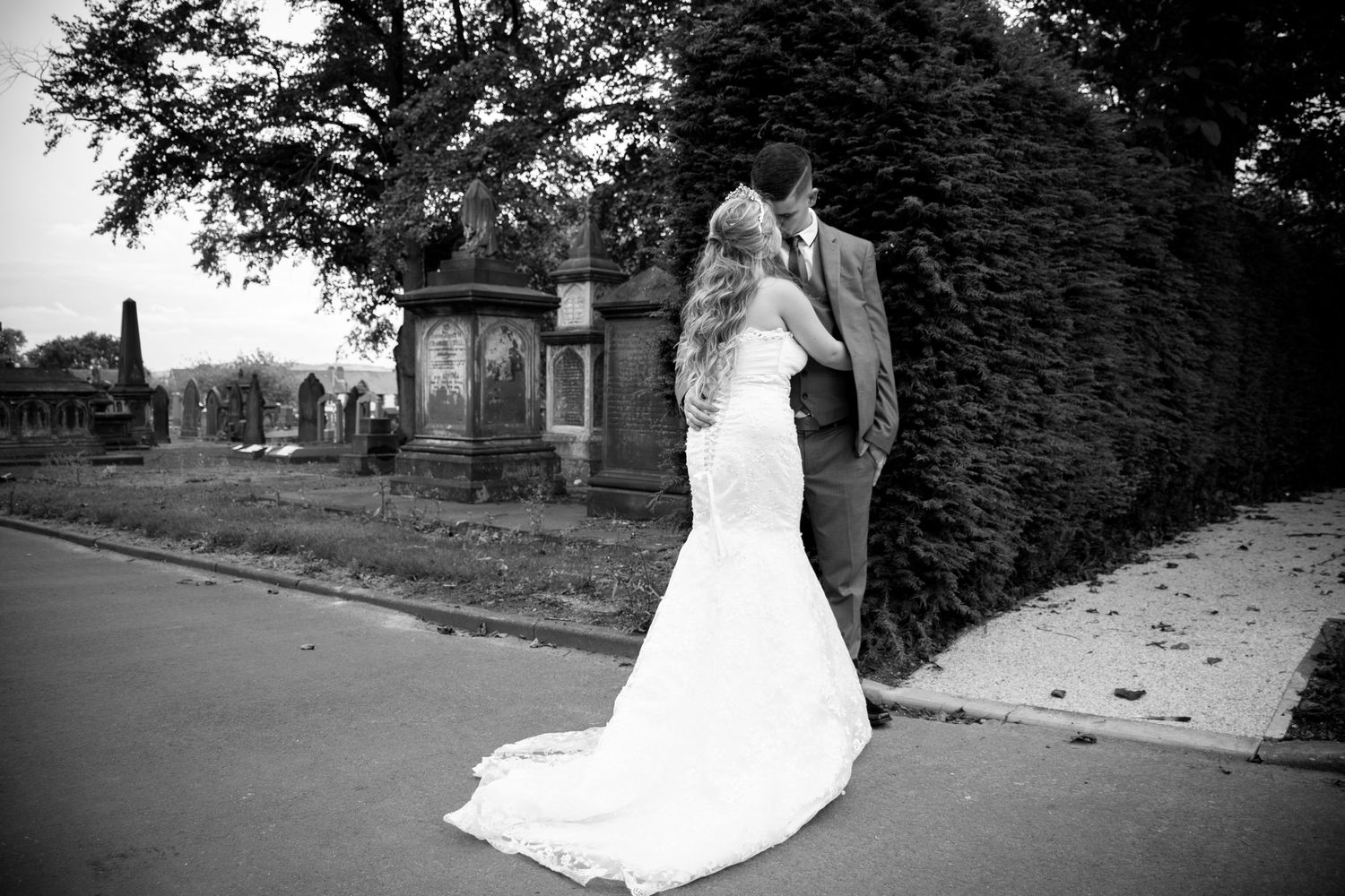 Black and white photo of a bride and groom standing together near a church hedge, with the bride's long train flowing behind her.