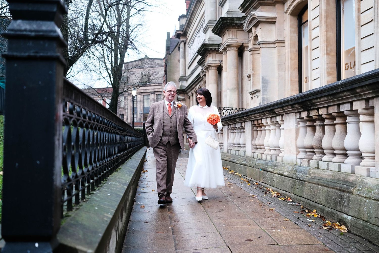 Bride and groom walking along a wet city pavement beside iron railings on a cloudy winter wedding day.