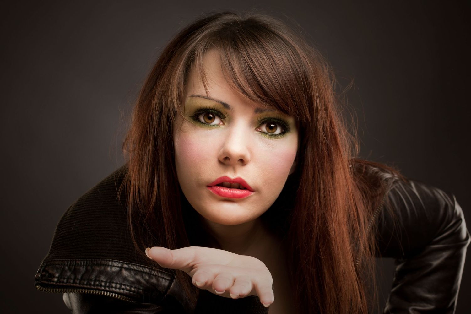 Young woman with long brown hair, green eye makeup, and red lips wearing a black leather jacket.