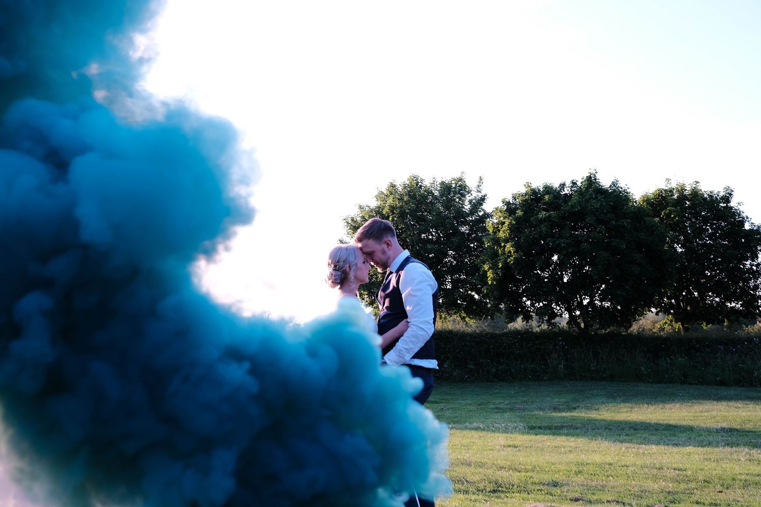 Couple sharing a romantic kiss surrounded by vibrant teal smoke bombs on a green field at their outdoor wedding.