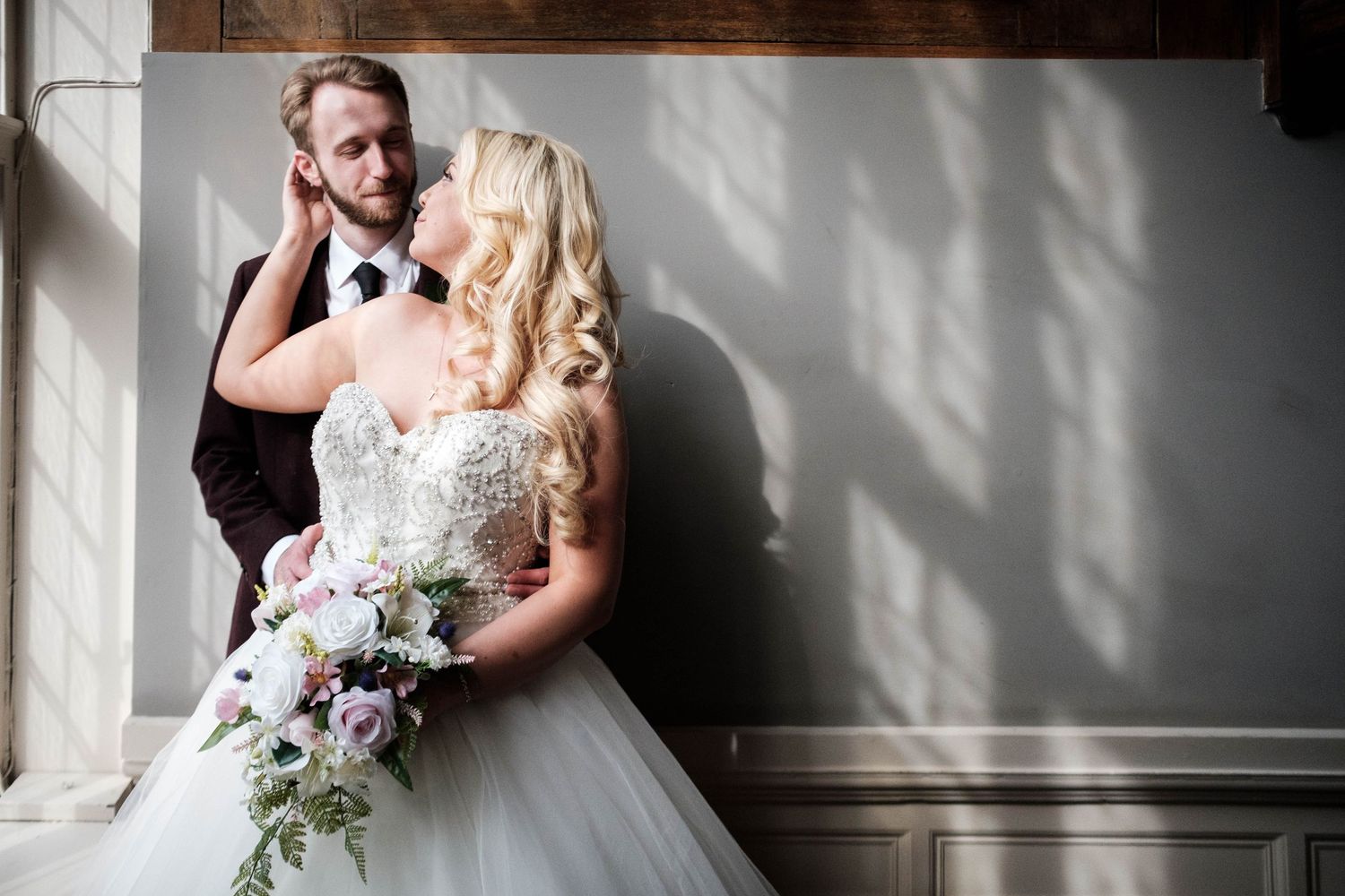 Bride in lace ballgown holding floral bouquet stands with groom in dark suit by sunlit window.