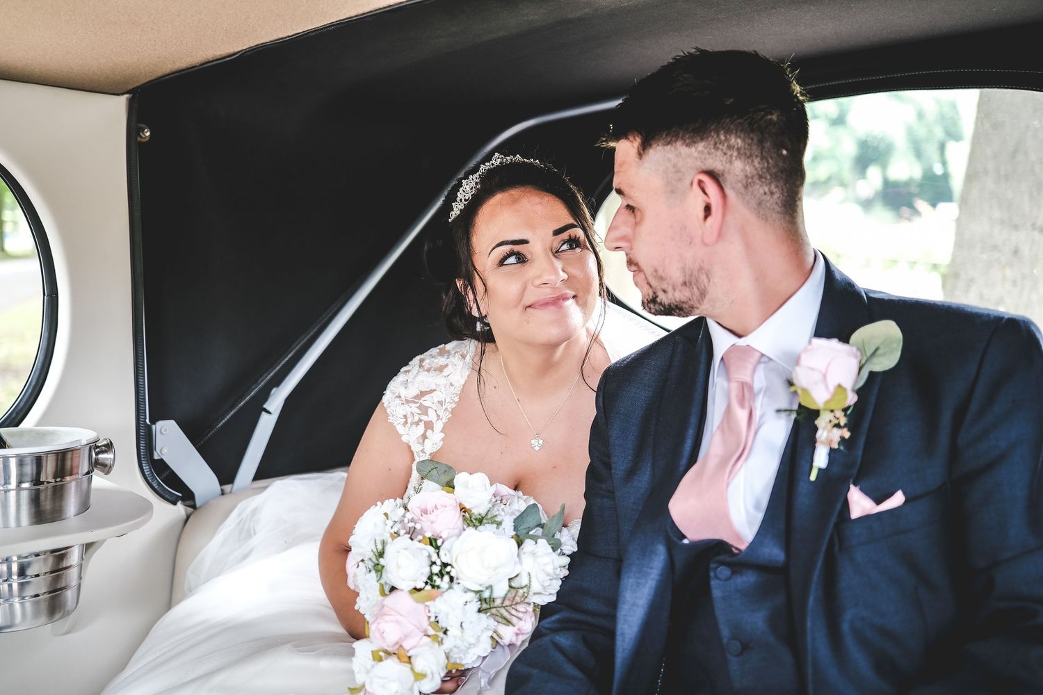 Bride and groom share a romantic moment inside a vintage wedding car, bride holding a white bouquet.