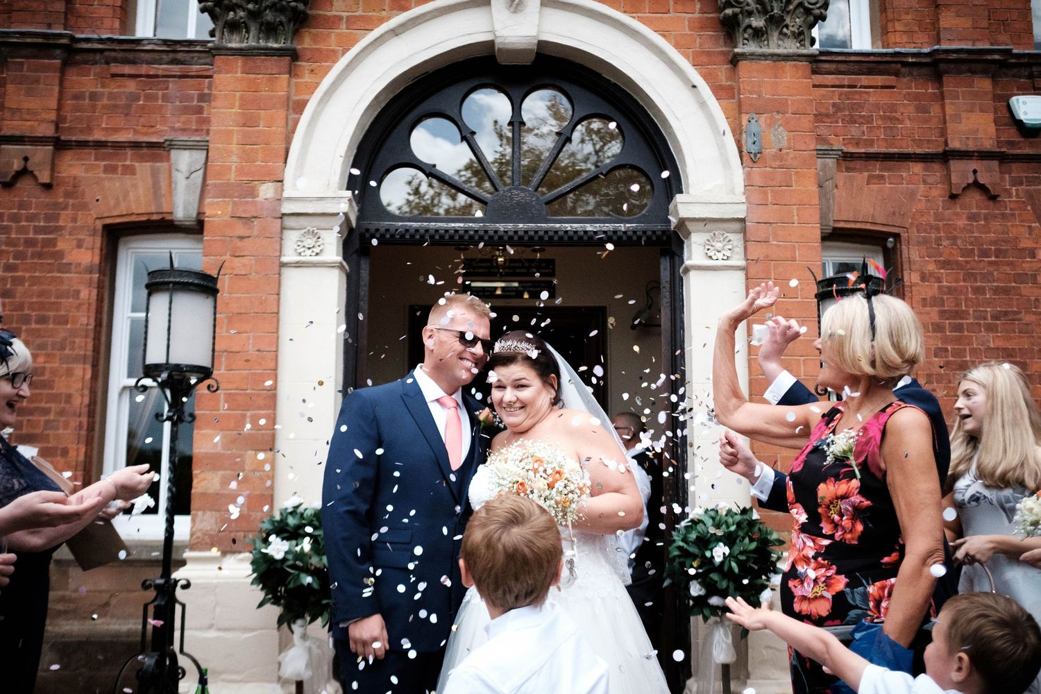 Bride and groom showered with confetti outside a red brick wedding venue surrounded by smiling guests.