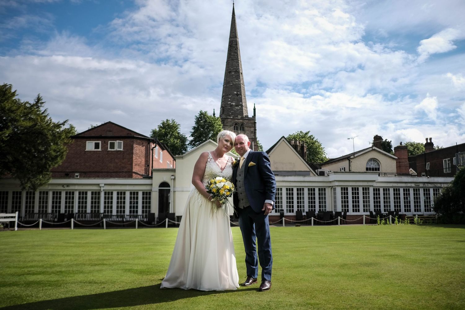 Bride and groom kissing on lawn with church spire and elegant venue in background on their wedding day.