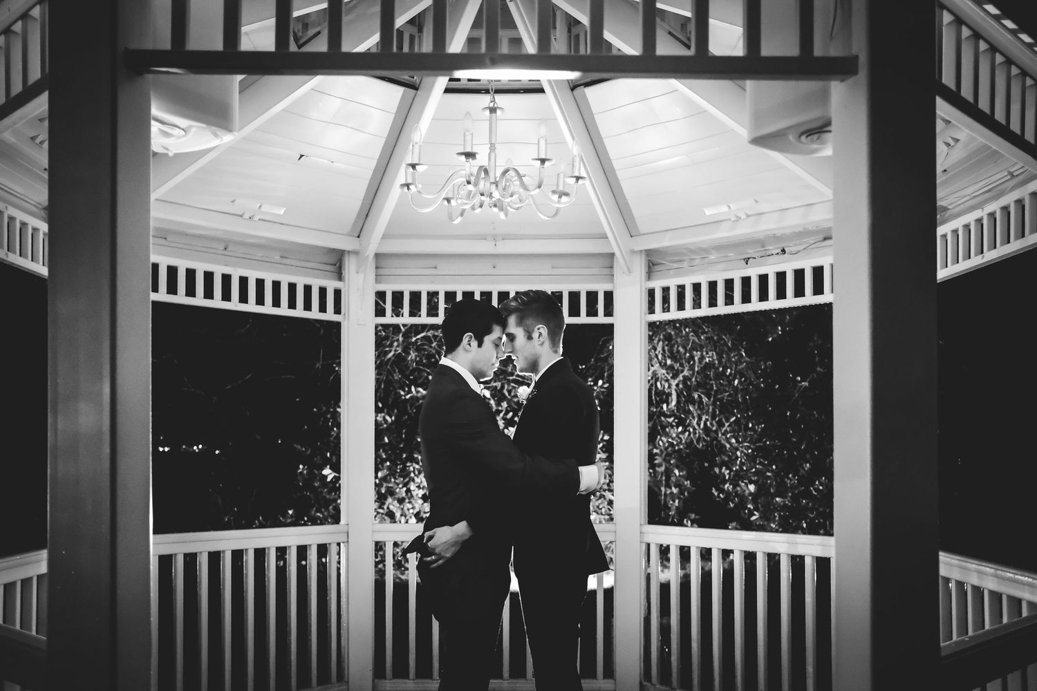 Two grooms share a romantic moment in a elegant gazebo with chandelier, captured in black and white wedding photography.
