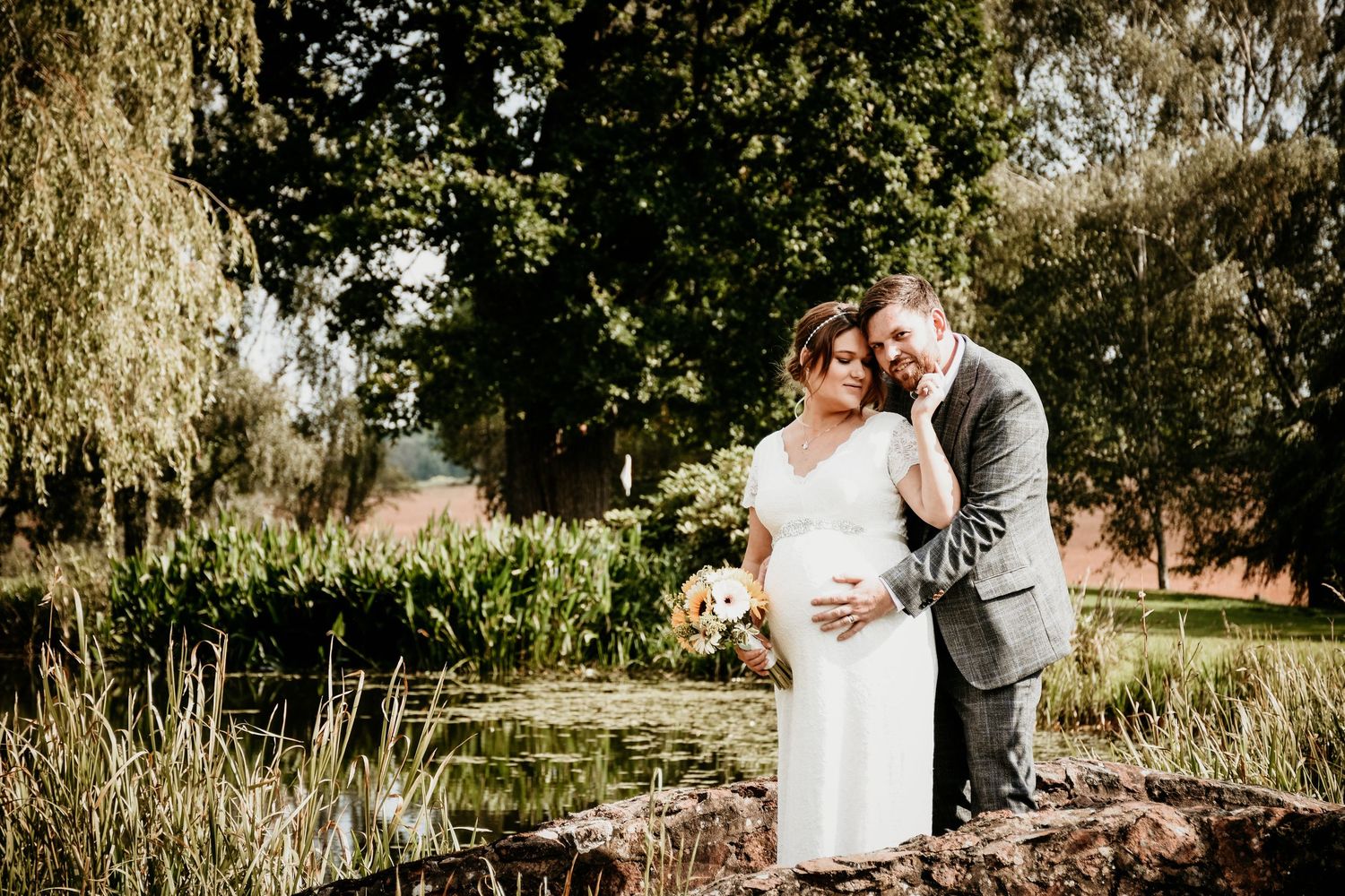 A couple poses by a pond in a lush garden, the bride in white holding flowers as the groom in gray embraces her.