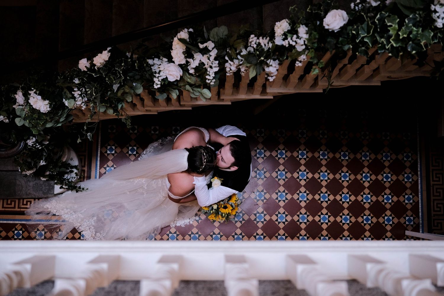Bride and groom share a romantic kiss near colorful tiled wall adorned with lush floral arrangements at wedding venue.