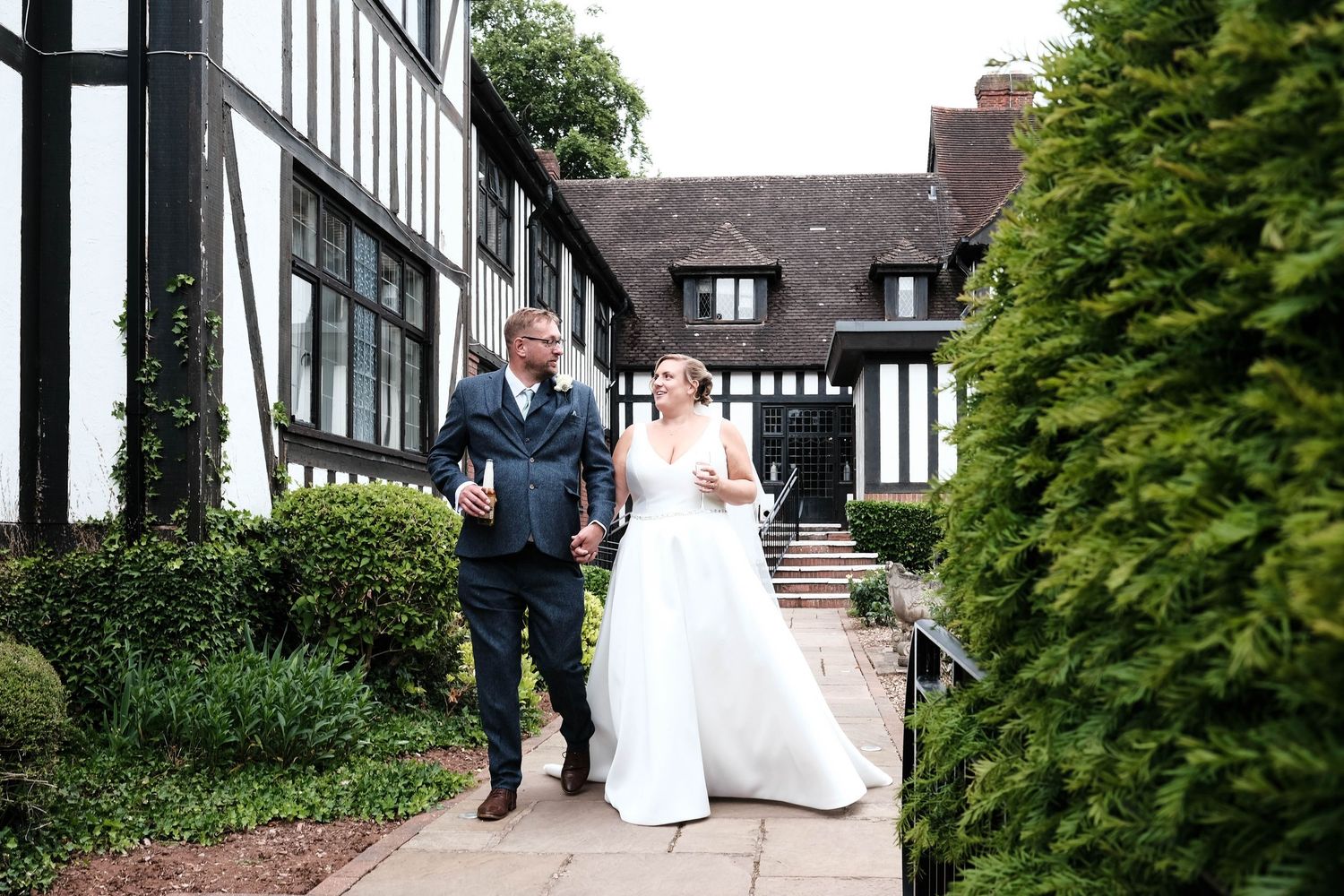 Bride and groom walk hand in hand outside a Tudor-style wedding venue surrounded by lush greenery.